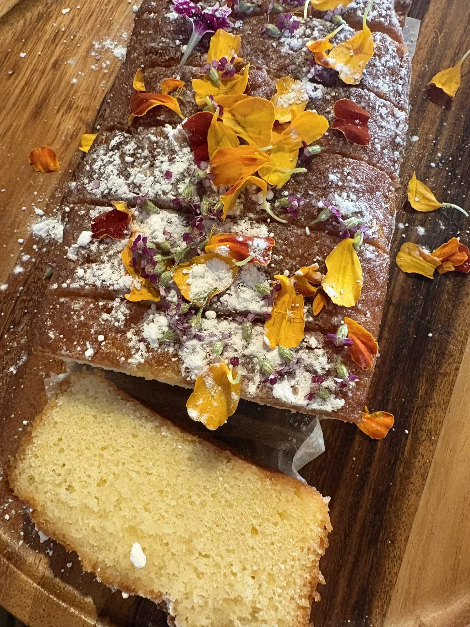 A piece of yellow cake on a wooden cutting board topped with powdered sugar, edible flowers, and purple microgreens.