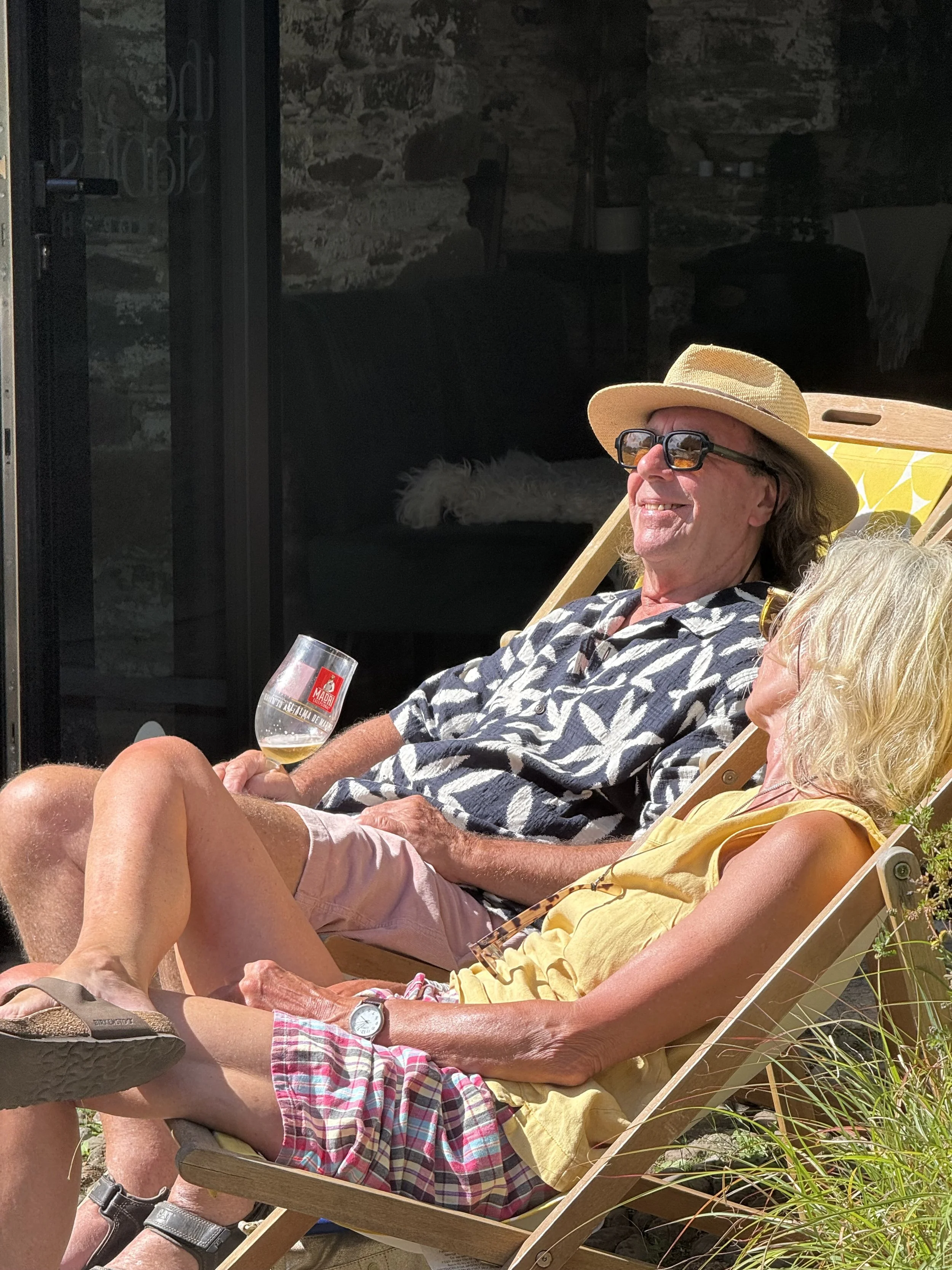 An elderly man and woman relaxing outdoors on a wooden lounge chair. The man is smiling, wearing a straw hat, sunglasses, a patterned short-sleeve shirt, and holding a glass of white wine. The woman has short, curly hair and is wearing a yellow top. 
