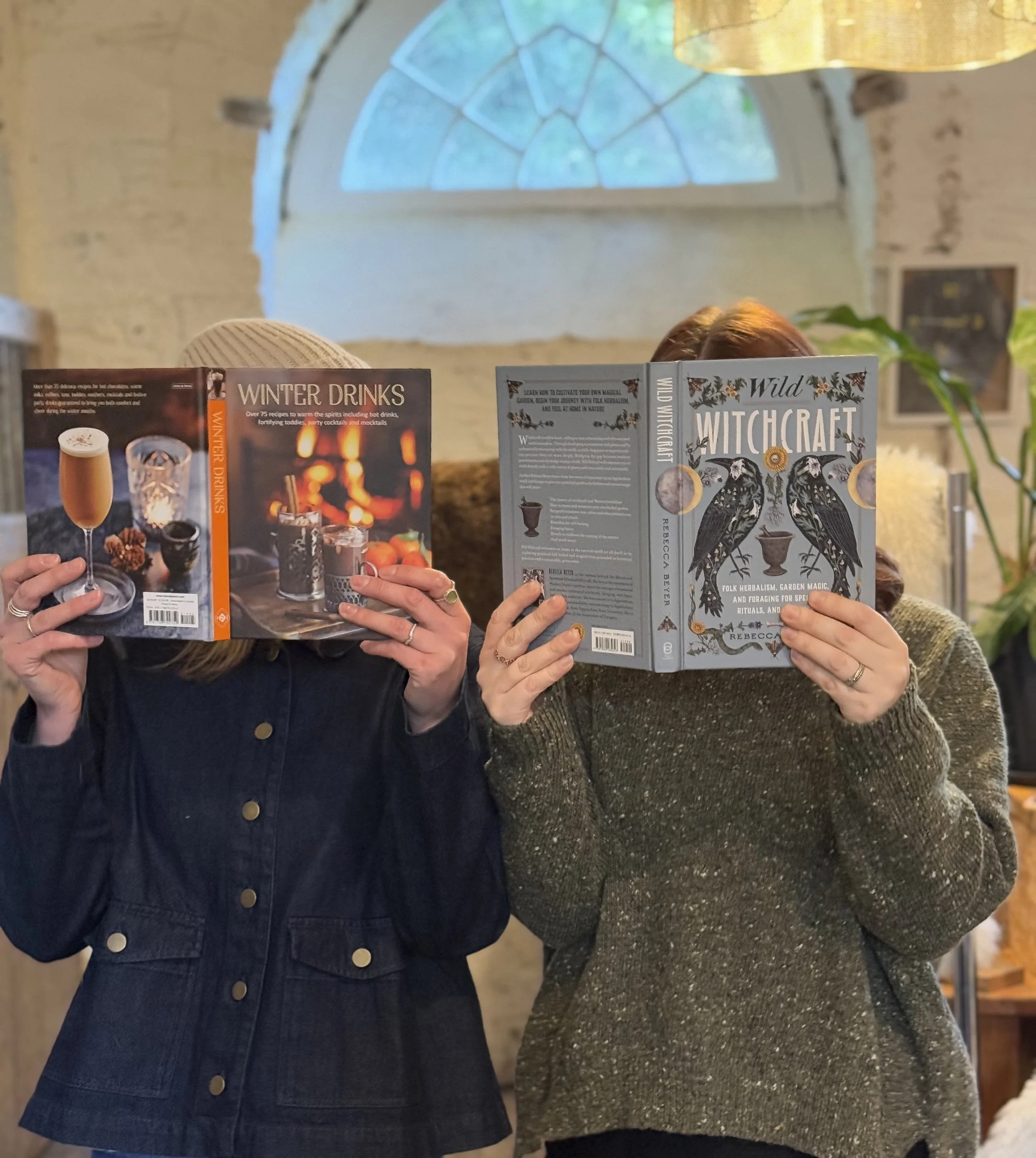 Two women holding open books or magazines, with a large arched window and a yellow hanging light in the background.