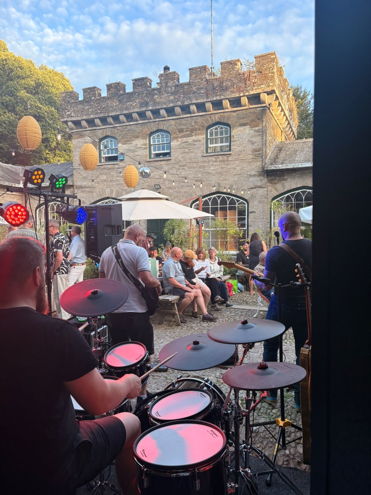 Outdoor concert or event with a band performing for an audience seated on benches. The scene is set in front of a stone building with arched windows, and decorative lanterns hang above. The sky is partly cloudy, and stage lights are visible on the le