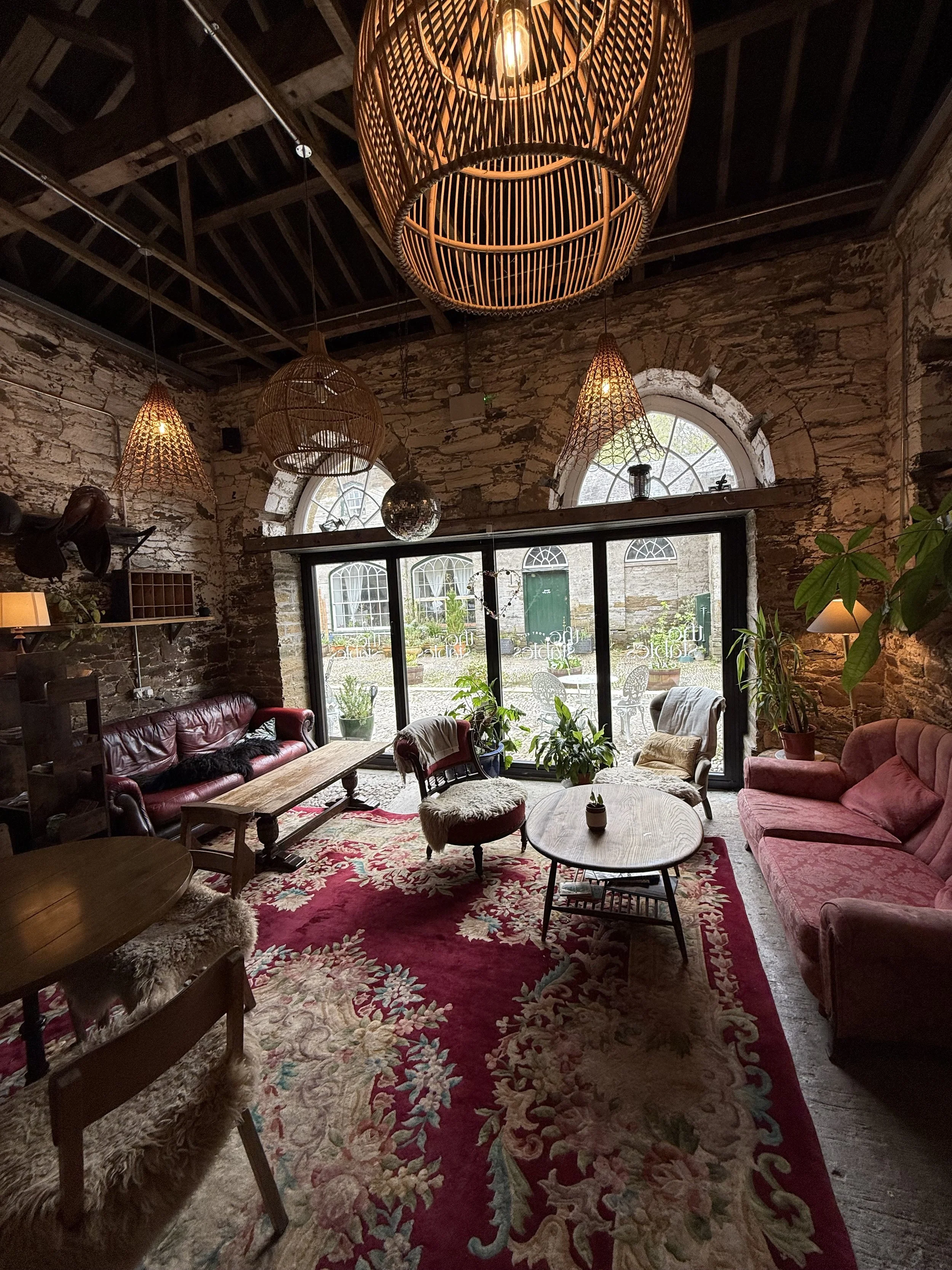 A cozy living room with exposed brick walls, large arched windows, and a high ceiling with wooden beams. The room features vintage-style furniture, including red velvet sofas, a round wooden coffee table, and patterned area rugs. There are potted pla