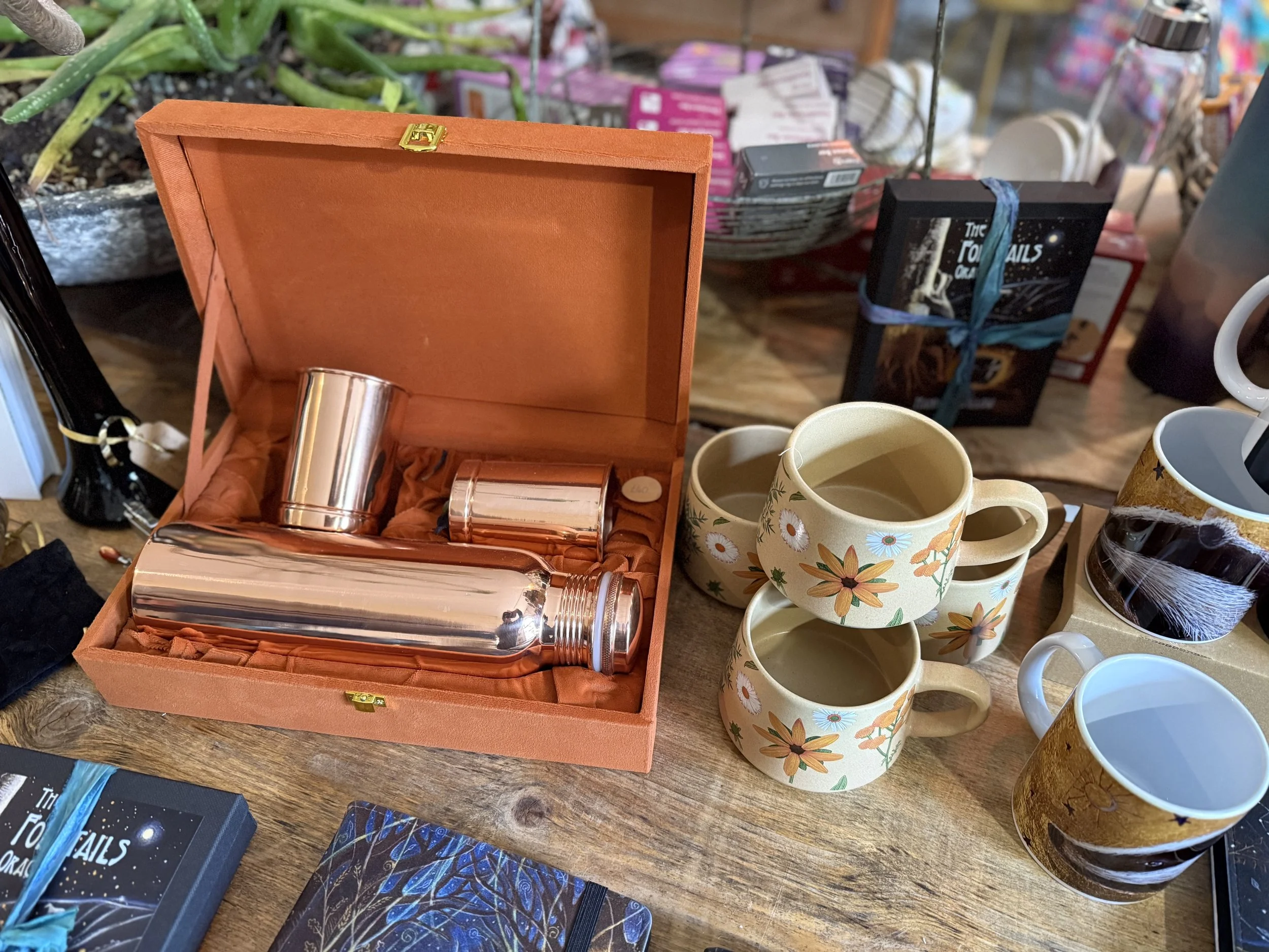 Various mugs with floral designs and a copper-colored stainless steel bottle inside an orange box, surrounded by other items like pens, books, and containers on a wooden surface.