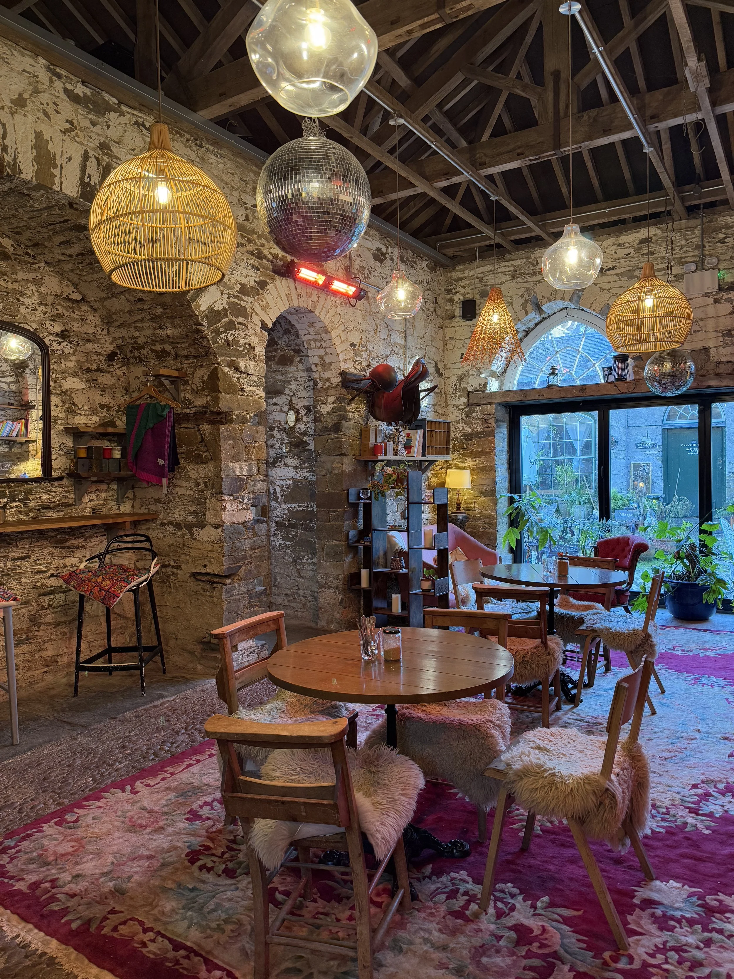 Interior of a cozy cafe or living space with exposed stone brick walls, high wooden ceiling, various hanging light fixtures including a disco ball, rattan pendant lamps, and a mix of modern and rustic furniture. There are plants, shelves with books, 