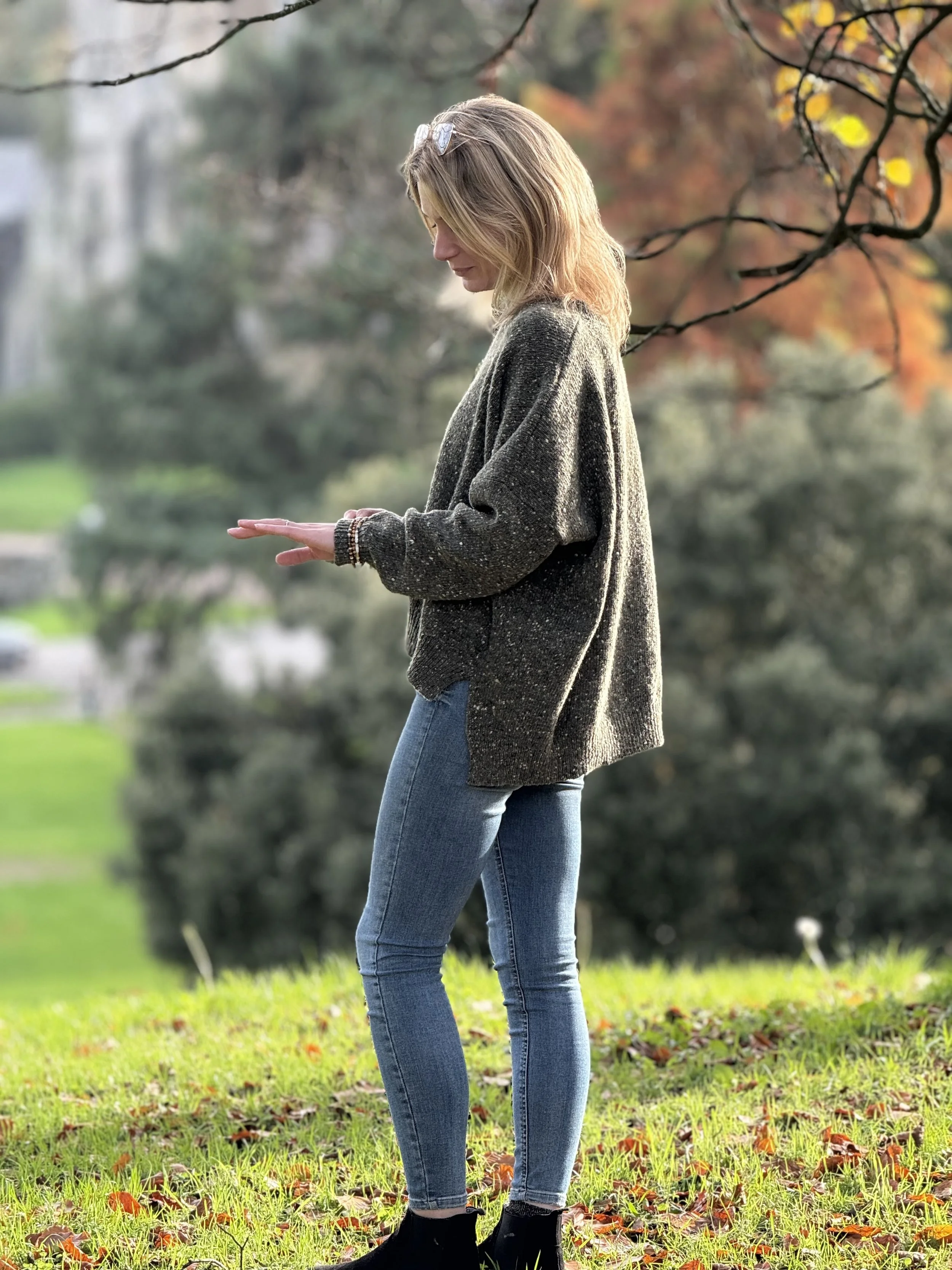 A woman with blonde hair in an outdoor park during autumn, wearing a gray sweater, blue jeans, black boots, and sunglasses on her head, standing on grass covered with fallen leaves, with trees and a park background.