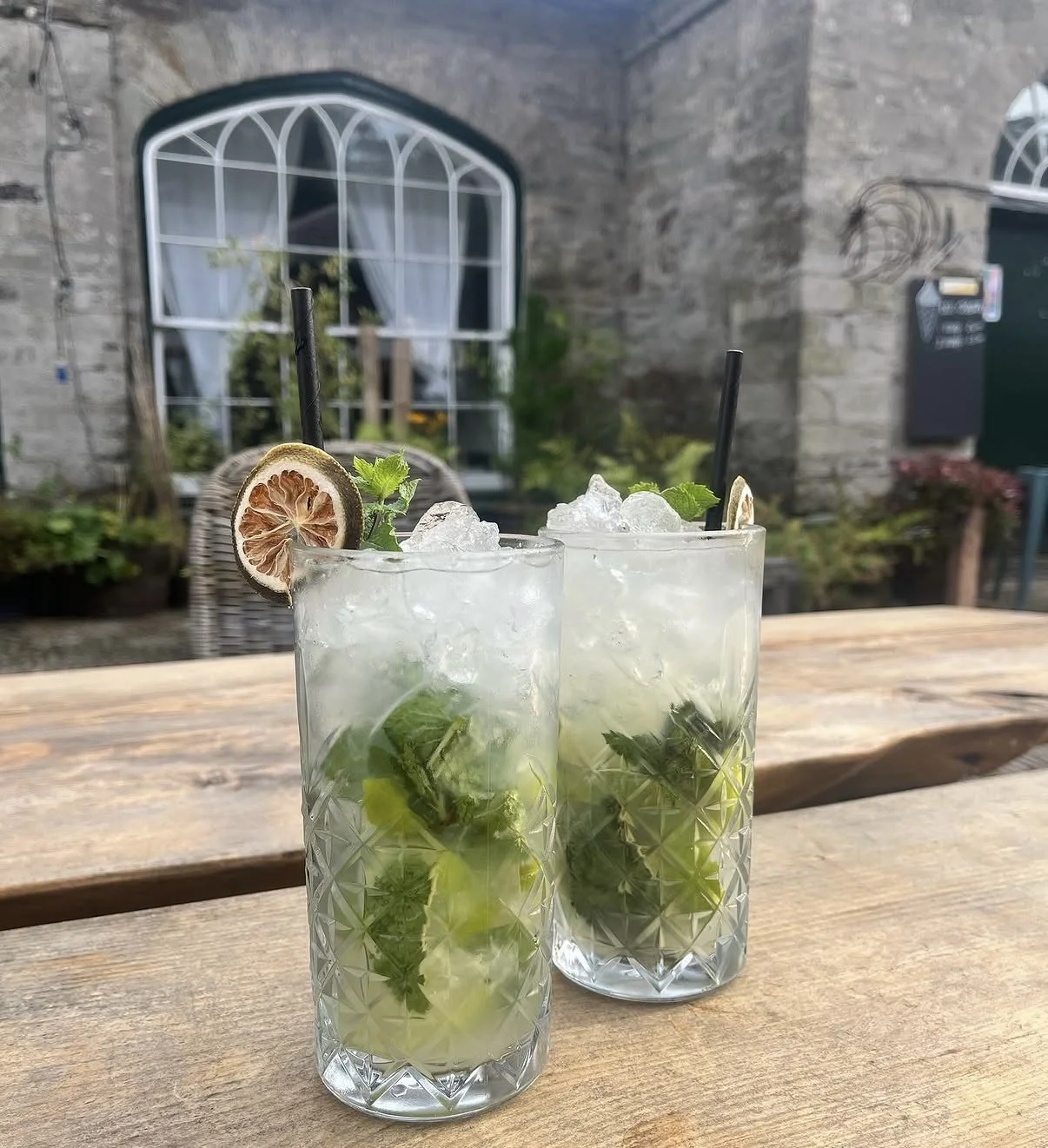 Two glasses of mojito cocktails with mint leaves, lime slices, and ice on a wooden table outdoors, with a rustic stone building and window in the background.