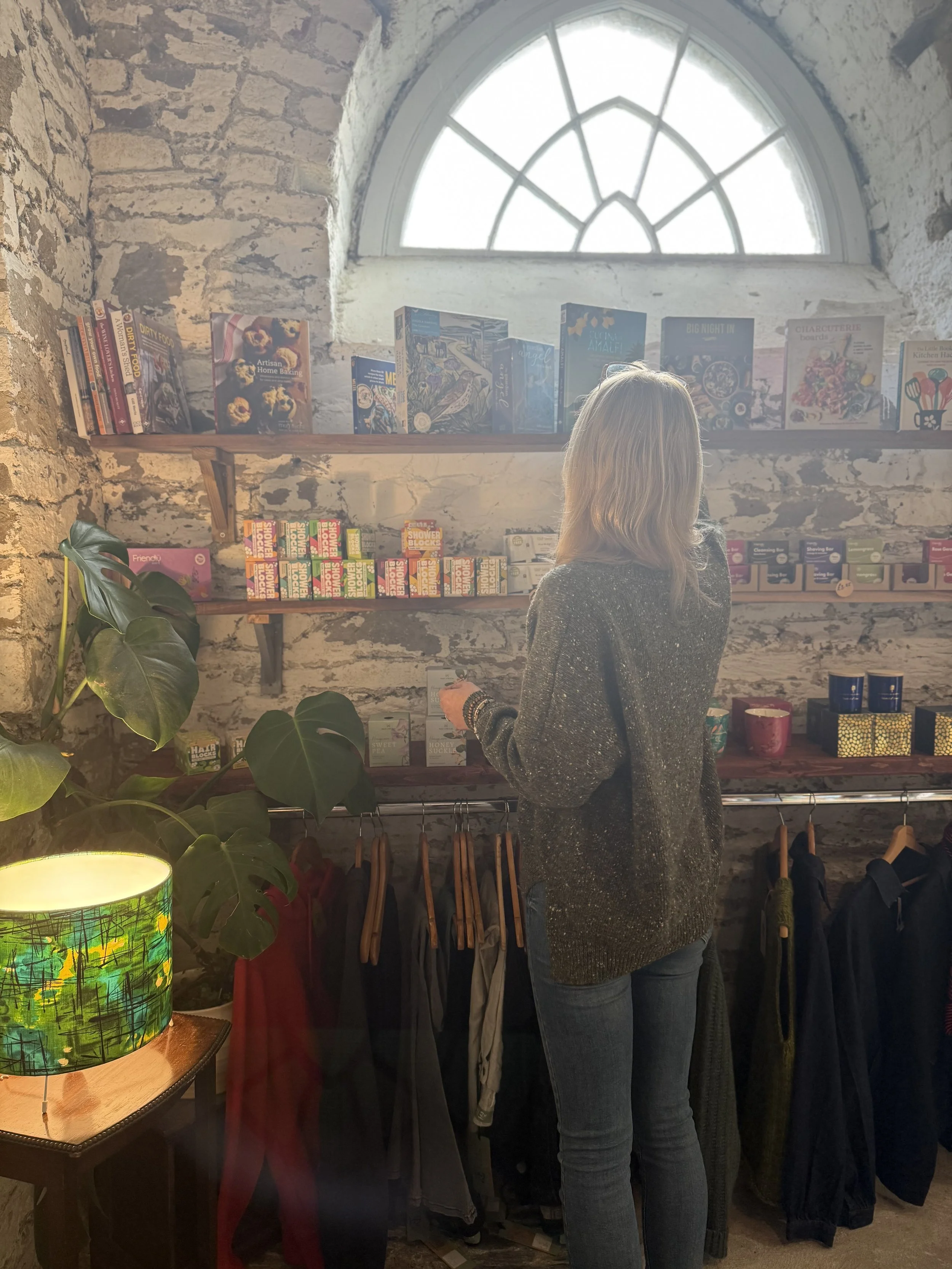 A woman shopping in a boutique store with rustic brick walls and wooden shelves. She is examining clothing on a rack with various coats and jackets. Behind her, a large arched window lets in natural light, illuminating a display of books and other it