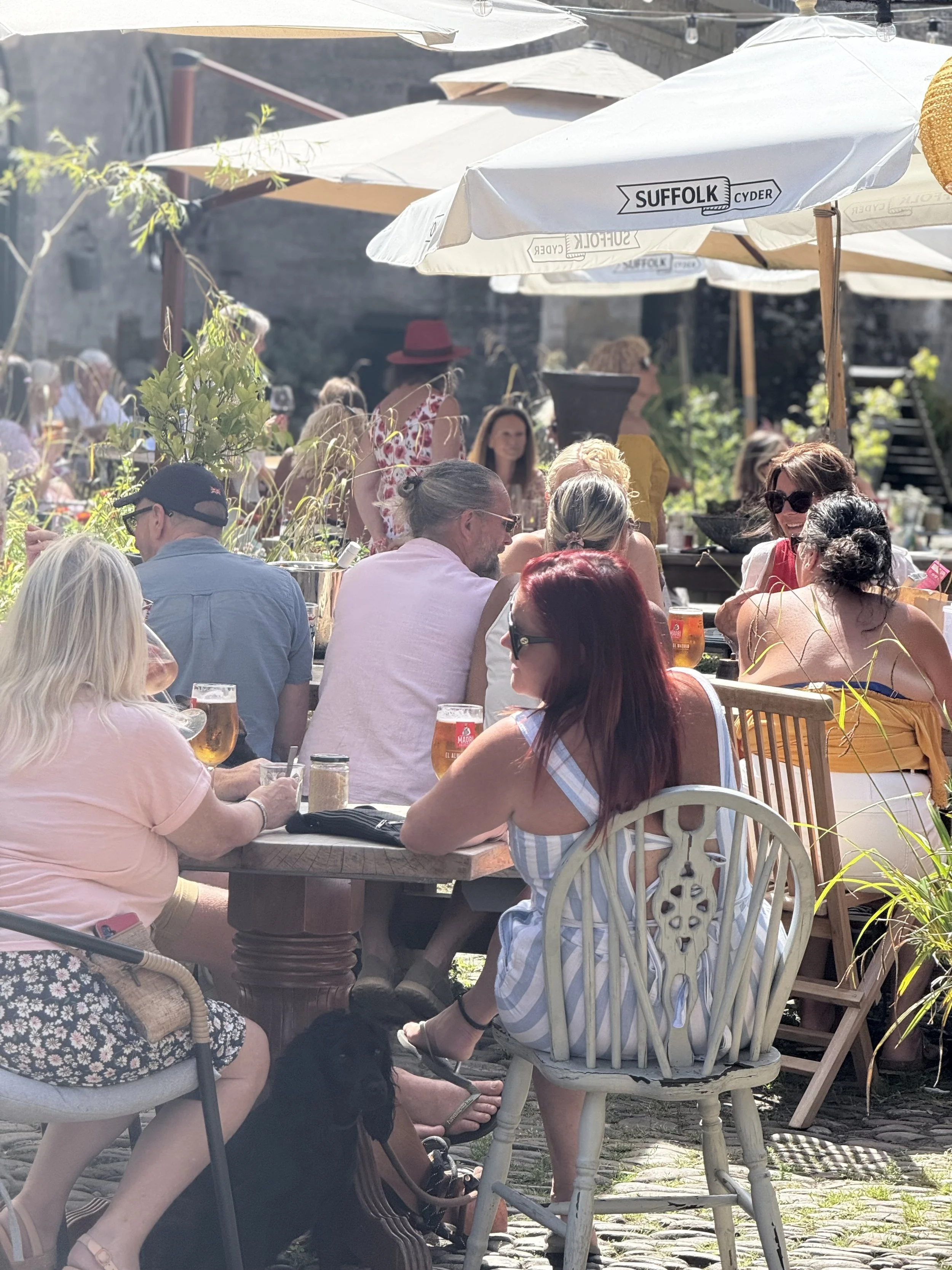 A group of people sitting at outdoor tables under umbrellas in a sunny setting, socializing and enjoying drinks.