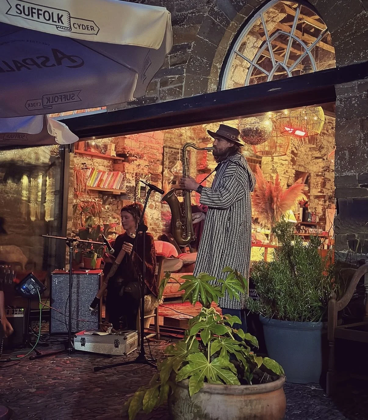 A nighttime scene at a cozy indoor venue with brick walls, illuminated by warm orange lighting and lanterns. A street musician wearing a striped coat and hat plays a saxophone, while a young woman with a microphone stands nearby. Potted plants and de