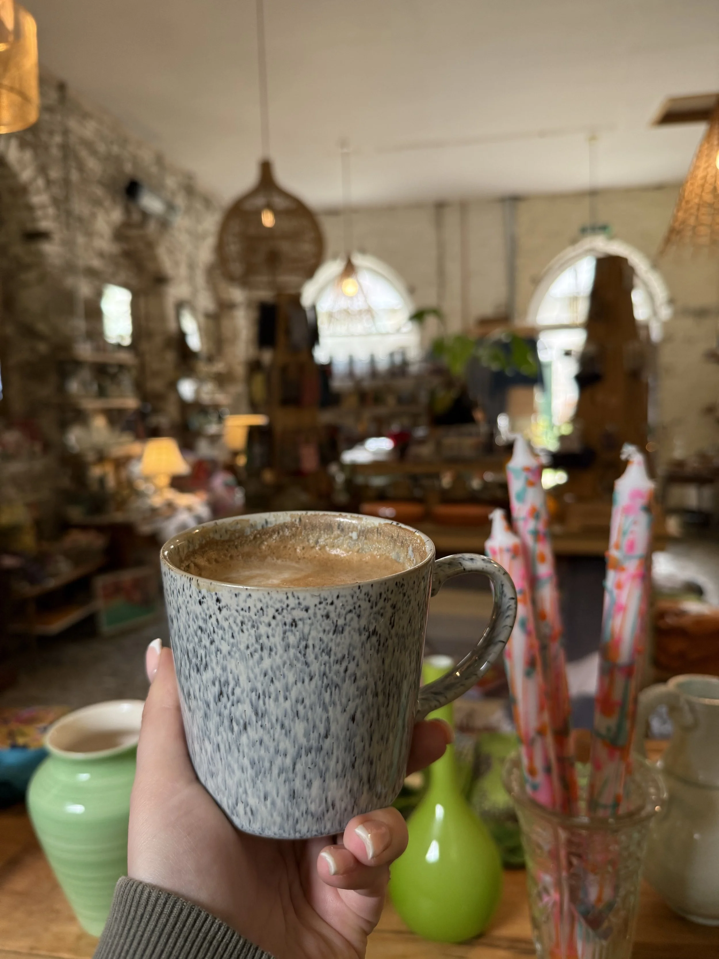 A hand holding a speckled ceramic mug filled with a hot beverage in a cozy store with vases and candles in the background.