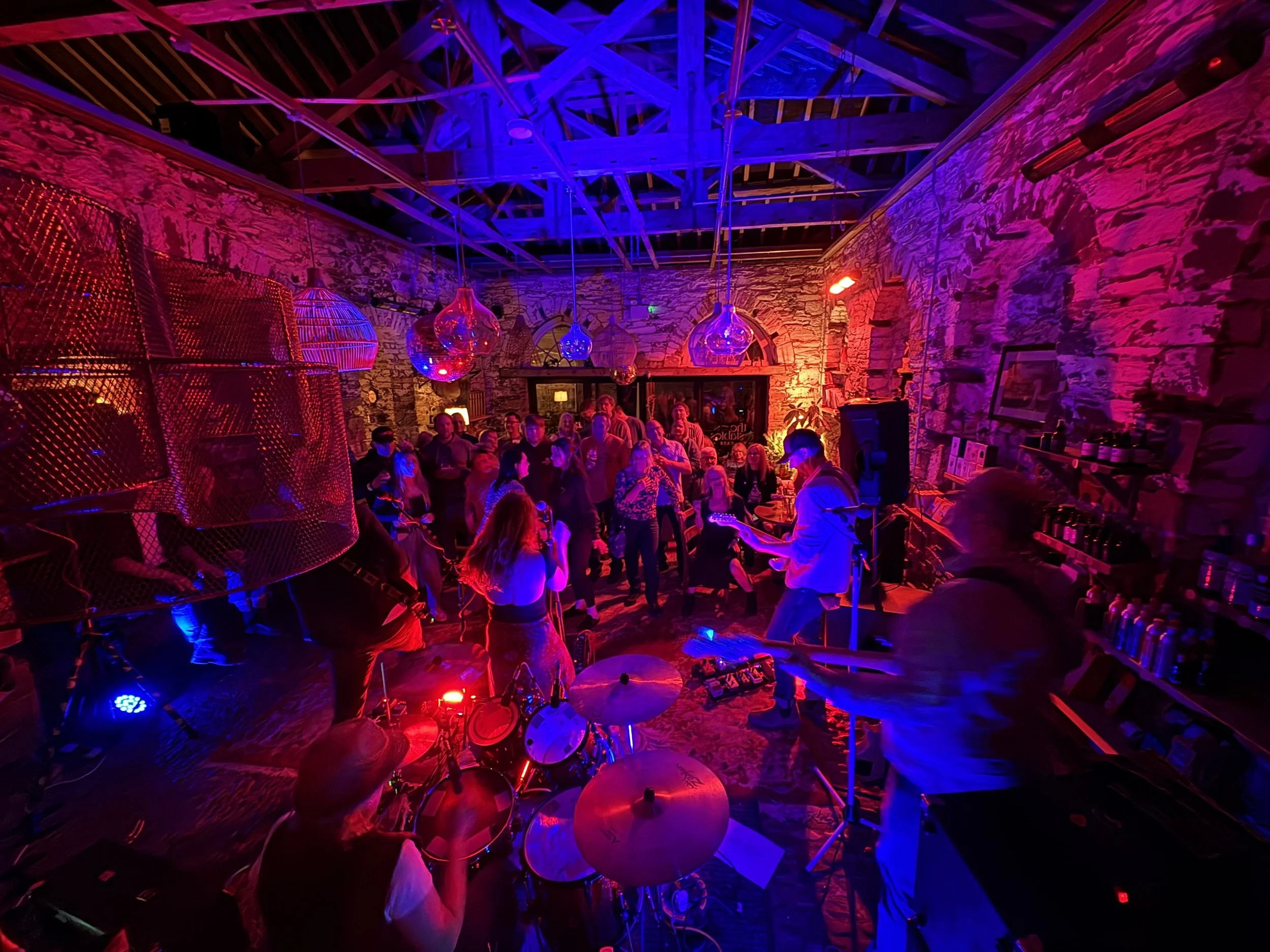 A band performing on stage with colorful blue and red lighting, in a venue with exposed brick walls and a wooden ceiling, with an audience watching.