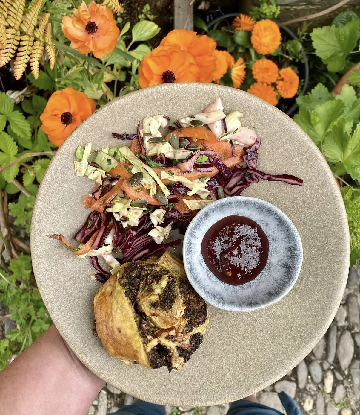 A plate of food held outdoors with orange flowers in the background. The plate contains a baked dish, a salad with shredded cabbage, carrots, mixed greens, and pumpkin seeds, and a small bowl of dark sauce or chutney.