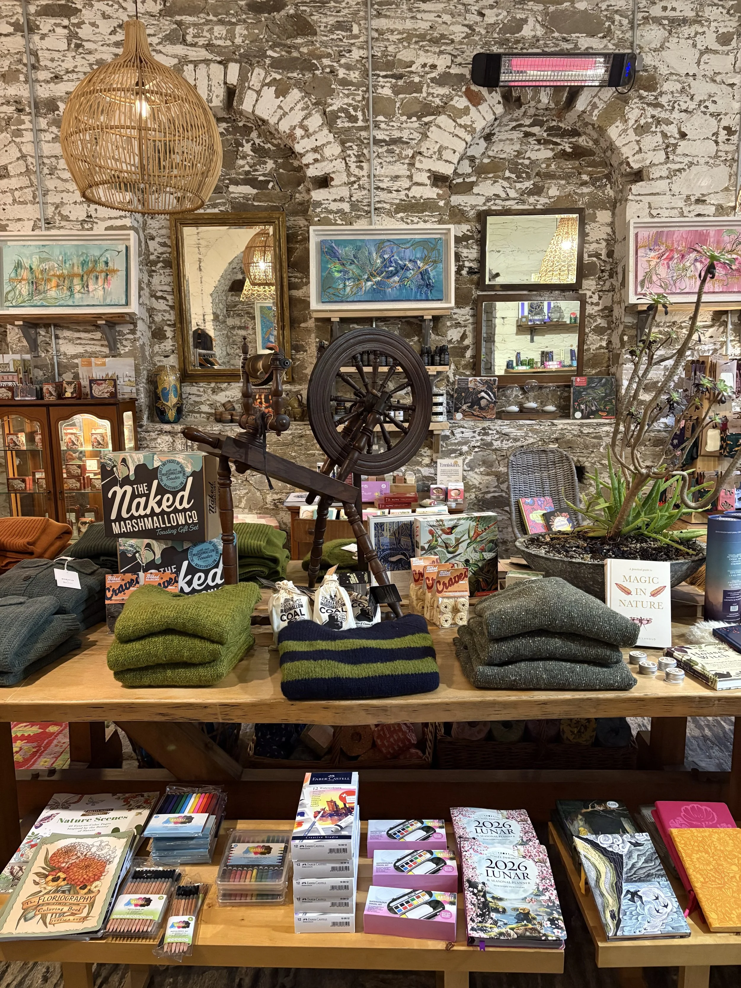 A display table with various colorful stationery, notebooks, and art supplies in a retail store with a rustic brick wall background. There are knitted hats and scarves on the table, and framed artwork on the wall behind, including paintings with abst