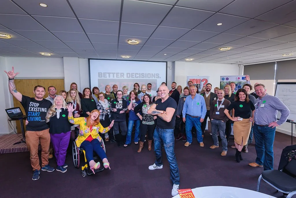 Group of diverse adults in a conference room, some smiling and waving, with a woman in a wheelchair in front. A large screen behind displays "BETTER DECISIONS."