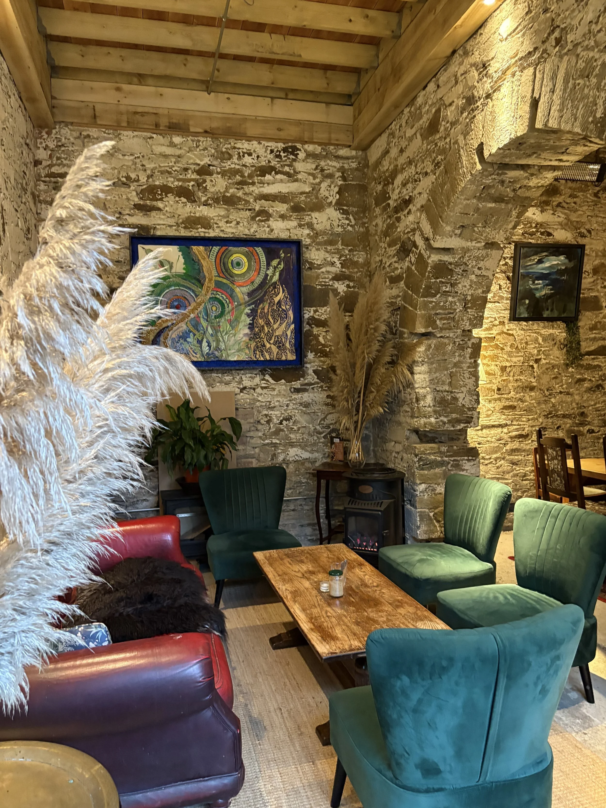 Cozy interior of a restaurant with a stone wall, colorful paintings, green and red chairs around a wooden table, a small electric fireplace, and tall pampas grass decorations.