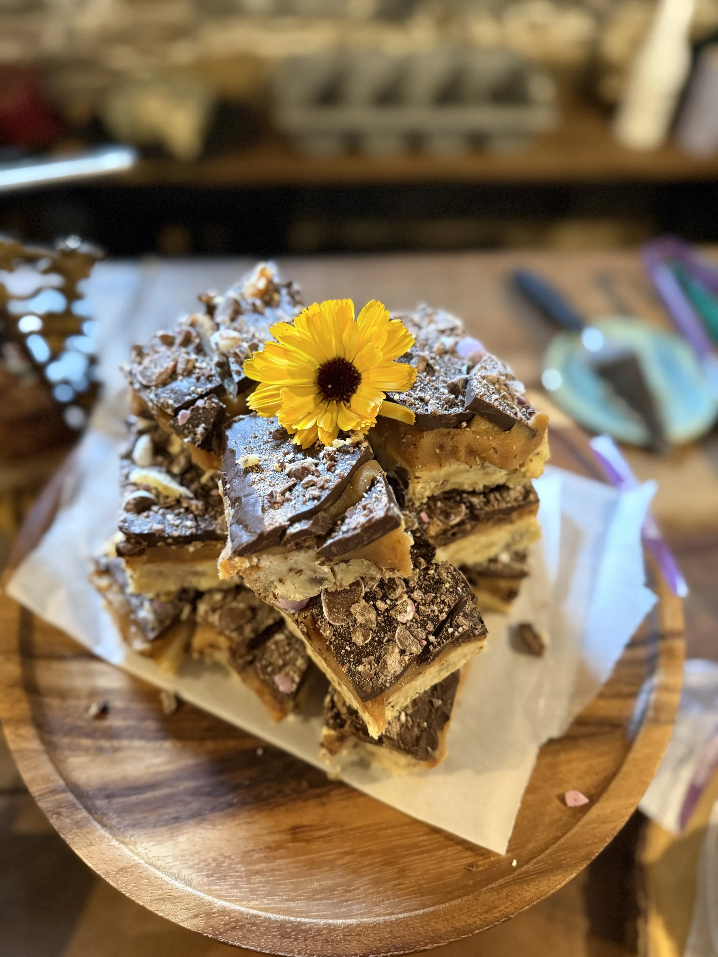 Stacked pieces of chocolate caramel shortbread with an edible yellow flower on top, on a round wooden serving tray.