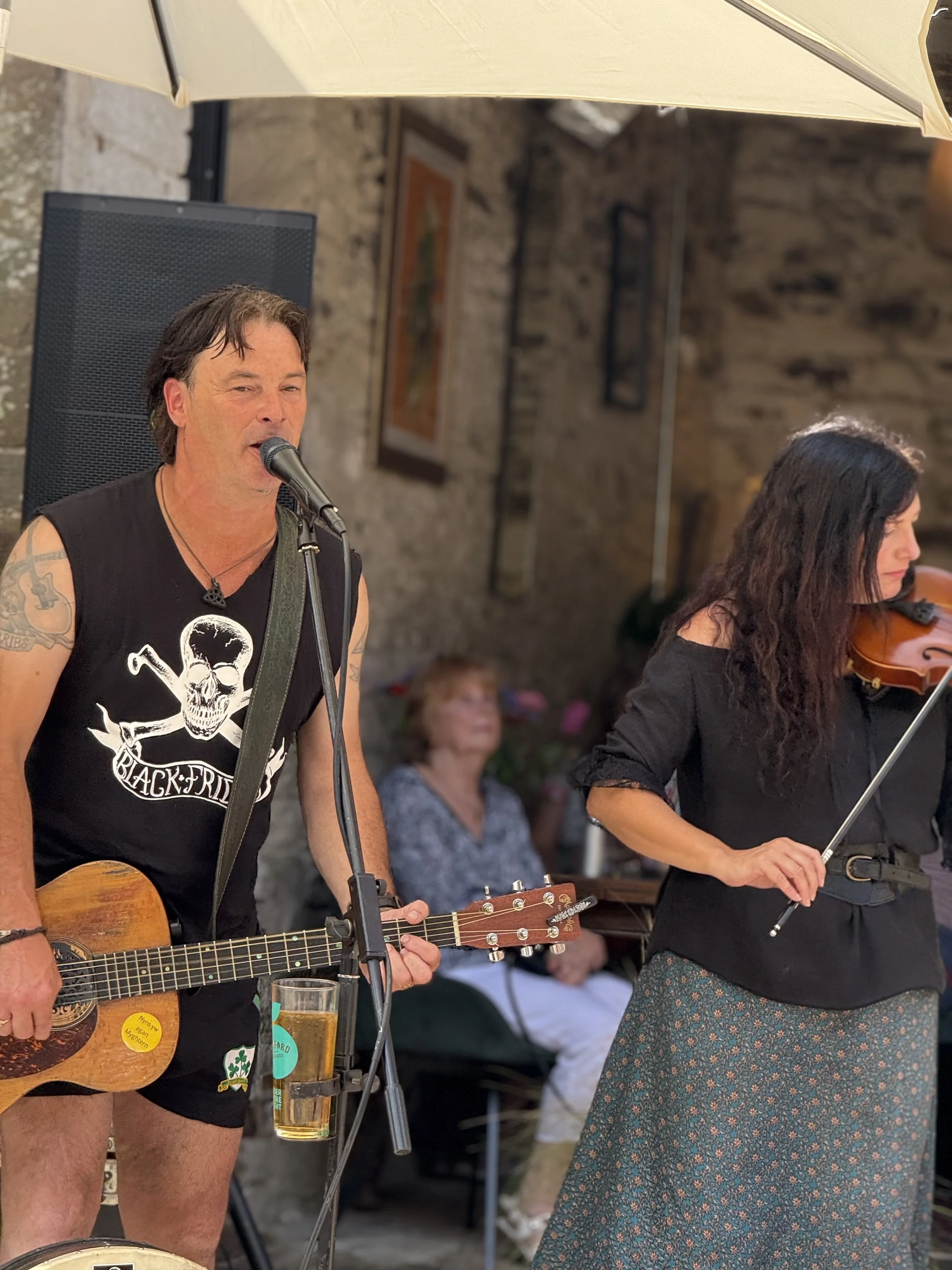 A man with dark hair, tattoos, and wearing a sleeveless black shirt with a skull design, playing an acoustic guitar and singing into a microphone at an outdoor event. A woman with long dark hair, wearing a black top and floral skirt, is playing a vio