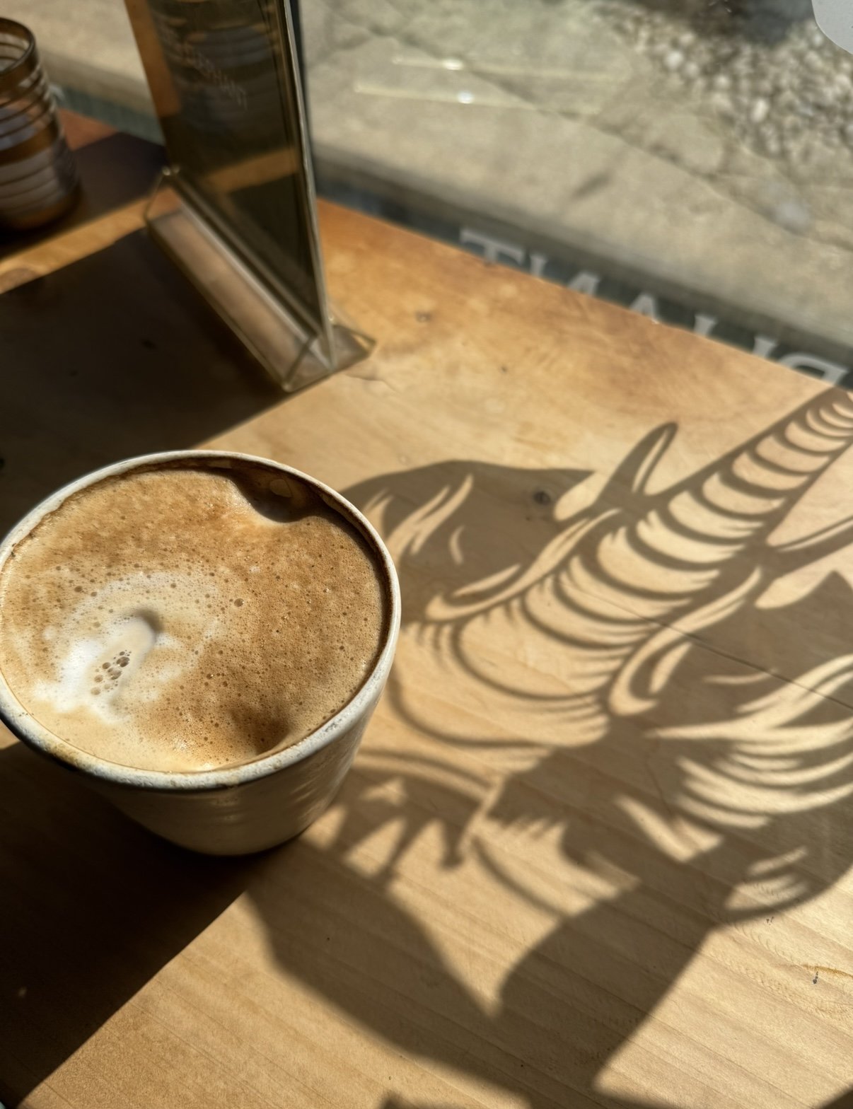 A cup of frothy coffee on a wooden table with intricate shadow patterns from a decorative light or object.