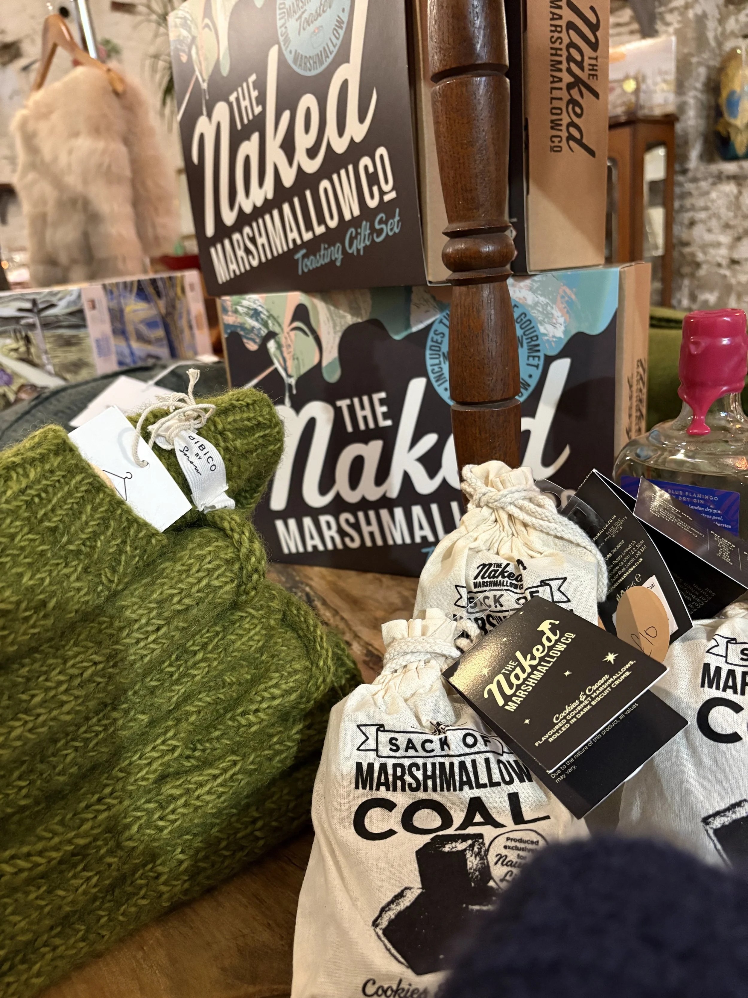 Close-up of a tabletop display with marshmallow-related products, including boxes of marshmallow treats, a pair of green knitted slippers, small fabric bags, and a glass jar with pink syrup, with a store background.