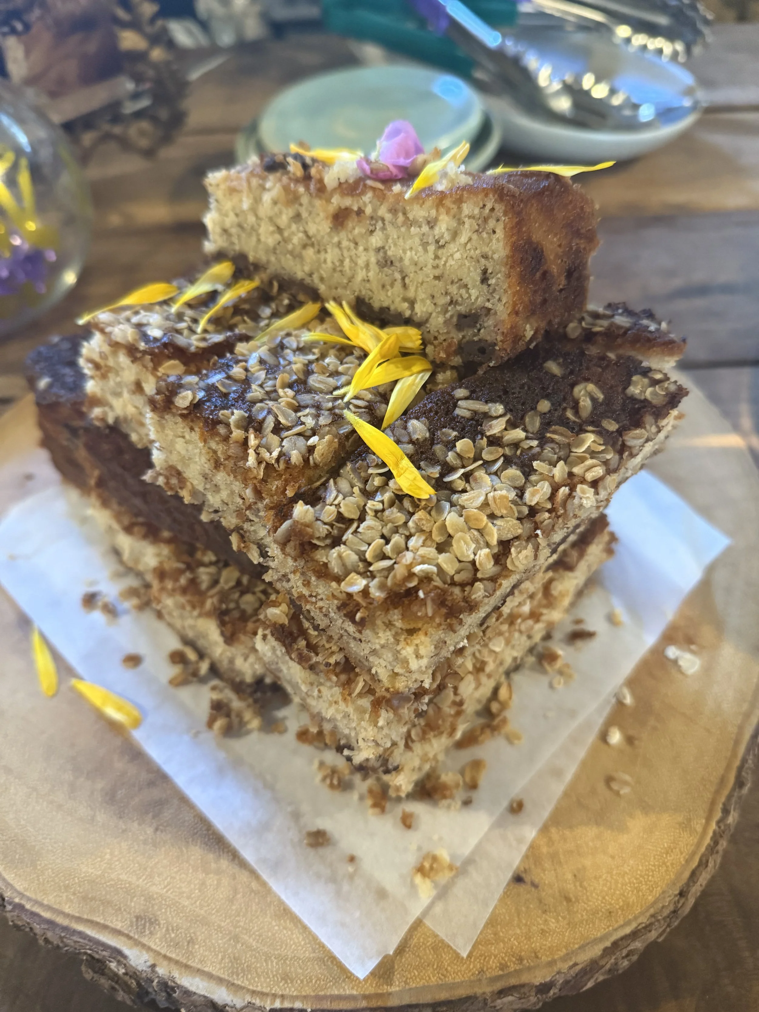 Close-up of a stack of three homemade crumb cake slices with chopped nuts on top, decorated with edible flower petals, placed on parchment paper on a wooden serving board.