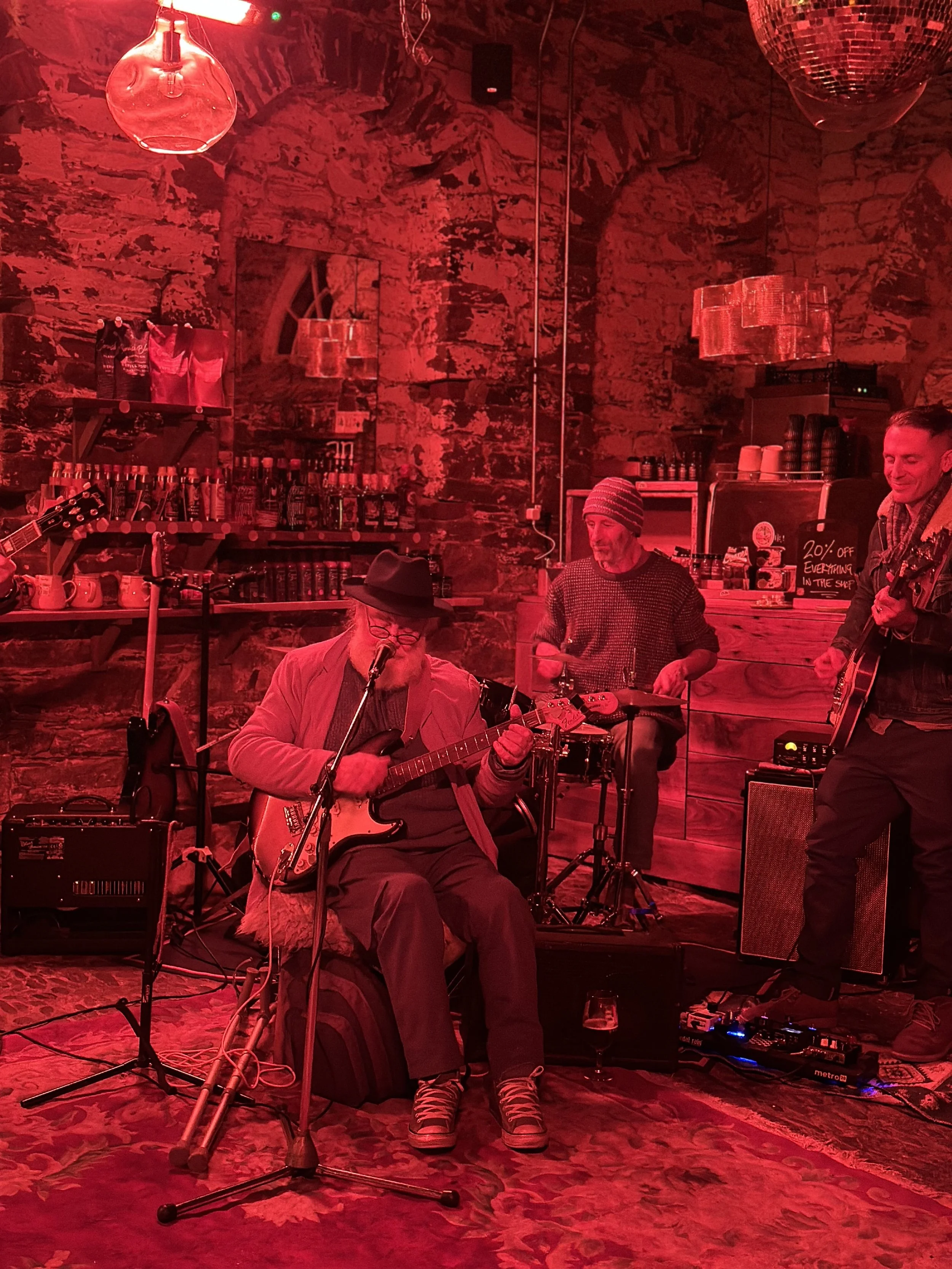 Three musicians performing on stage with red lighting. One man playing an electric guitar, another on drums, and a third with a guitar. The stage has brick walls, shelves with bottles, and a sign offering a discount.
