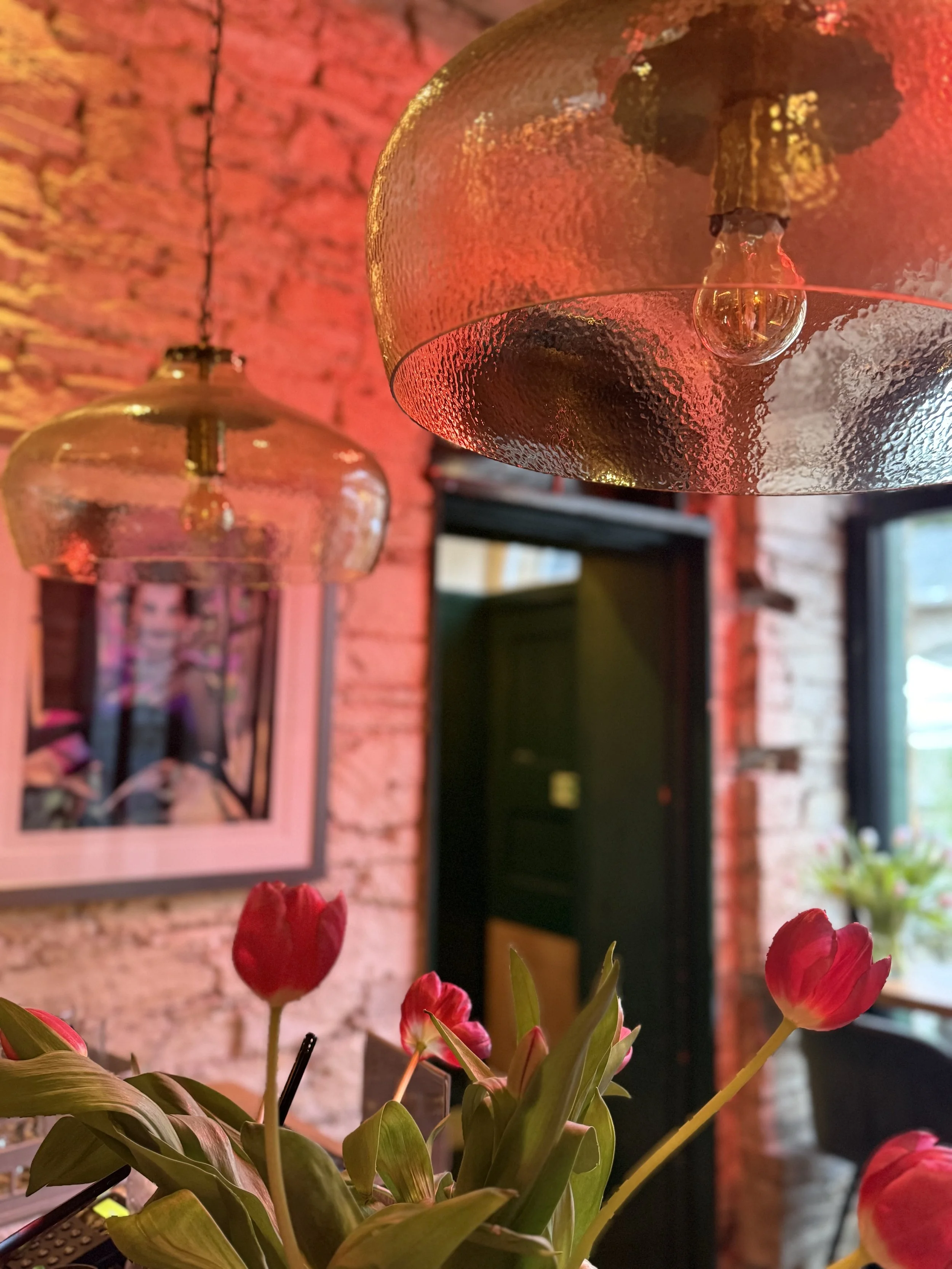 Close-up of red tulips with green leaves in the foreground. The background features a textured brick wall, a black shelf, a framed picture, and hanging lamps emitting a warm glow.