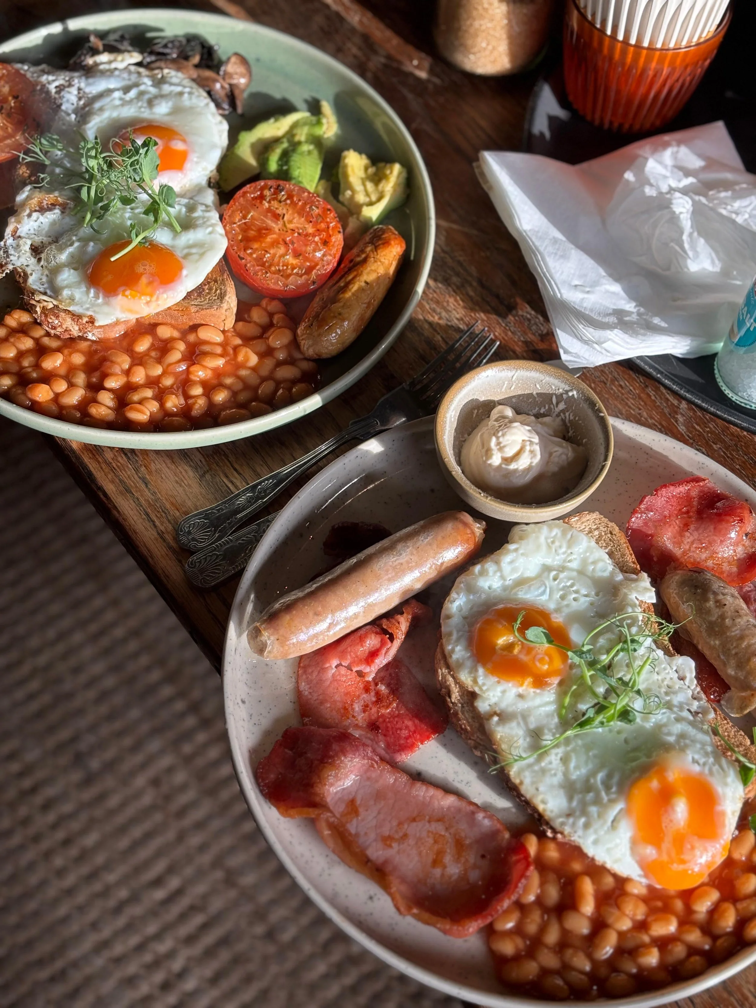 A breakfast plate with fried eggs, baked beans, avocado slices, tomato, sausage, bacon, toast, and a small cup of cream or yogurt. There is also a second plate with similar items and condiment jars in the background.