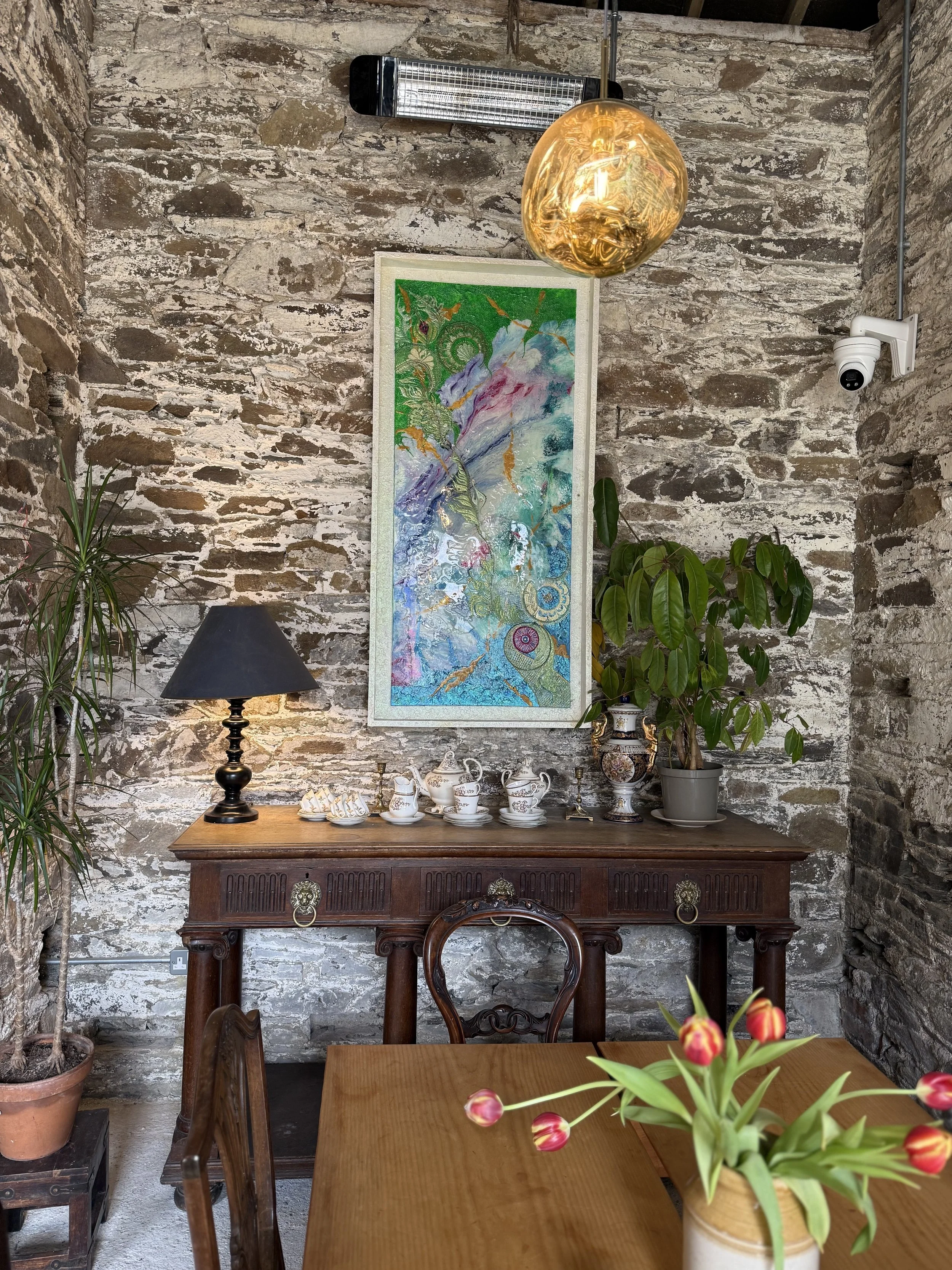 A rustic interior with exposed stone walls, a wooden sideboard displaying fine china, and plants. A modern gold spherical pendant light hangs above, complementing a sleek black table lamp. A colorful abstract painting is hung on the wall.
