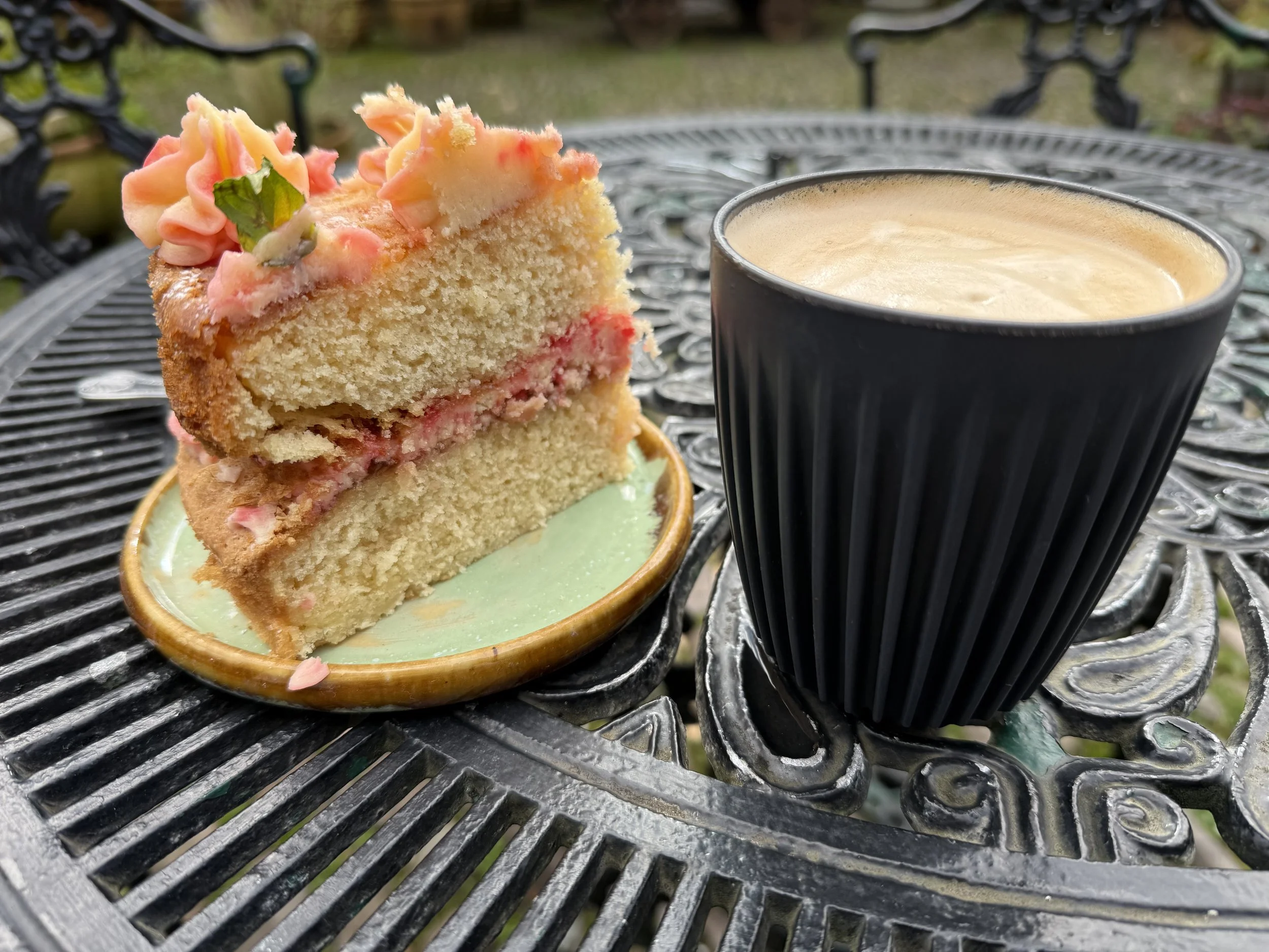 A slice of vanilla cake with pink frosting and green sprigs, served on a small green plate, next to a black mug of coffee with foam, on an outdoor black metal table.