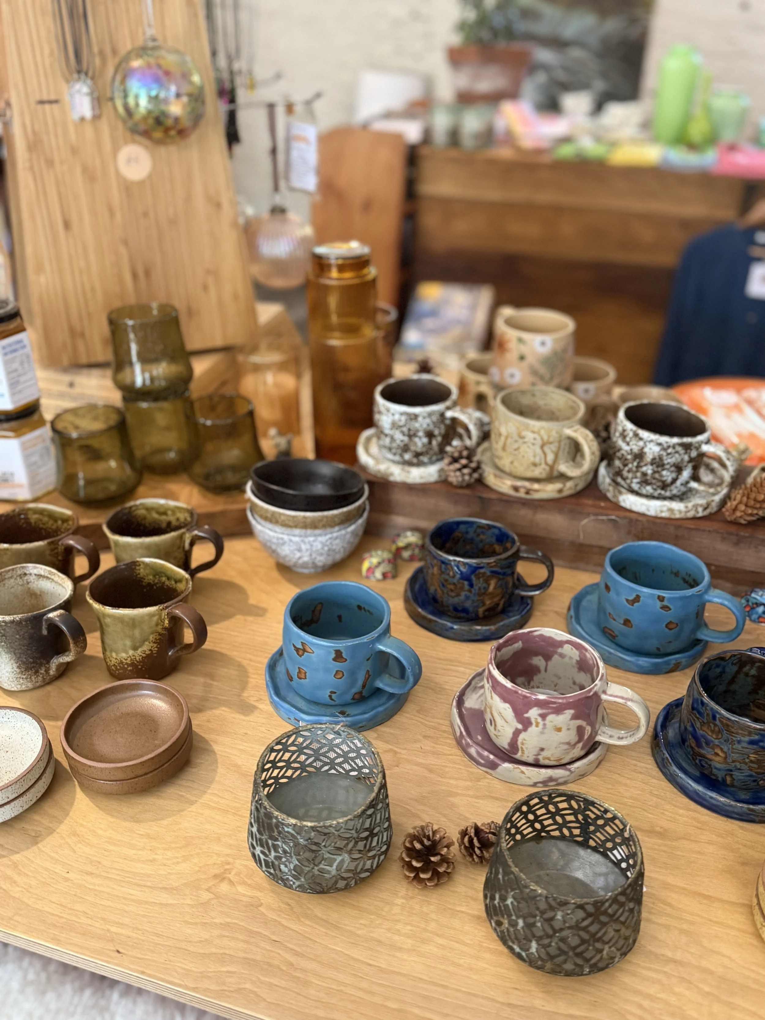 A display of various handcrafted ceramic mugs and cups in multiple colors and patterns on a wooden table, with pinecones and decorative metal candle holders in the foreground.