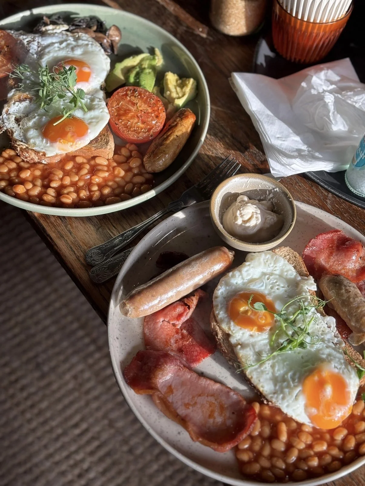 Two plates of breakfast with fried eggs, baked beans, sausage, bacon, avocado, tomato, and toast, on a wooden table.