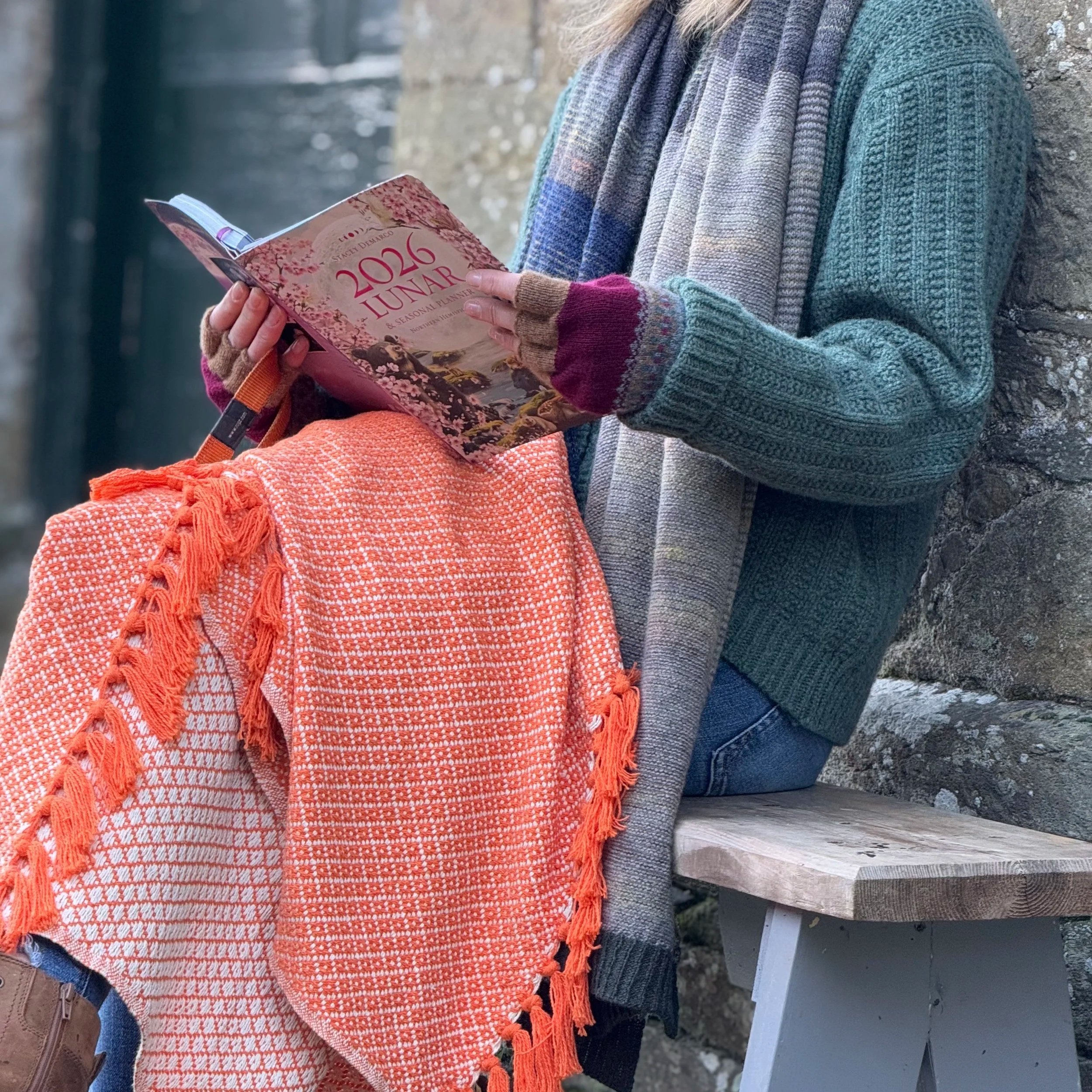 A person leaning against a stone wall, wearing a multi-colored scarf, a colorful knitted glove, and holding a 2026 lunar seasonal planner notebook. The person is also wearing a gray sweater and blue jeans, and is sitting beside a gray wooden bench.