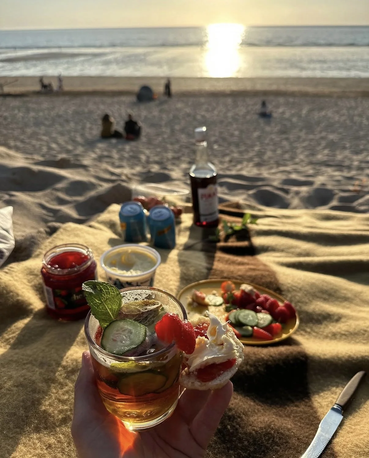 Beach picnic setup with a drink topped with mint and strawberries, assorted snacks, and condiments on a blanket with a sunset over the ocean in the background.