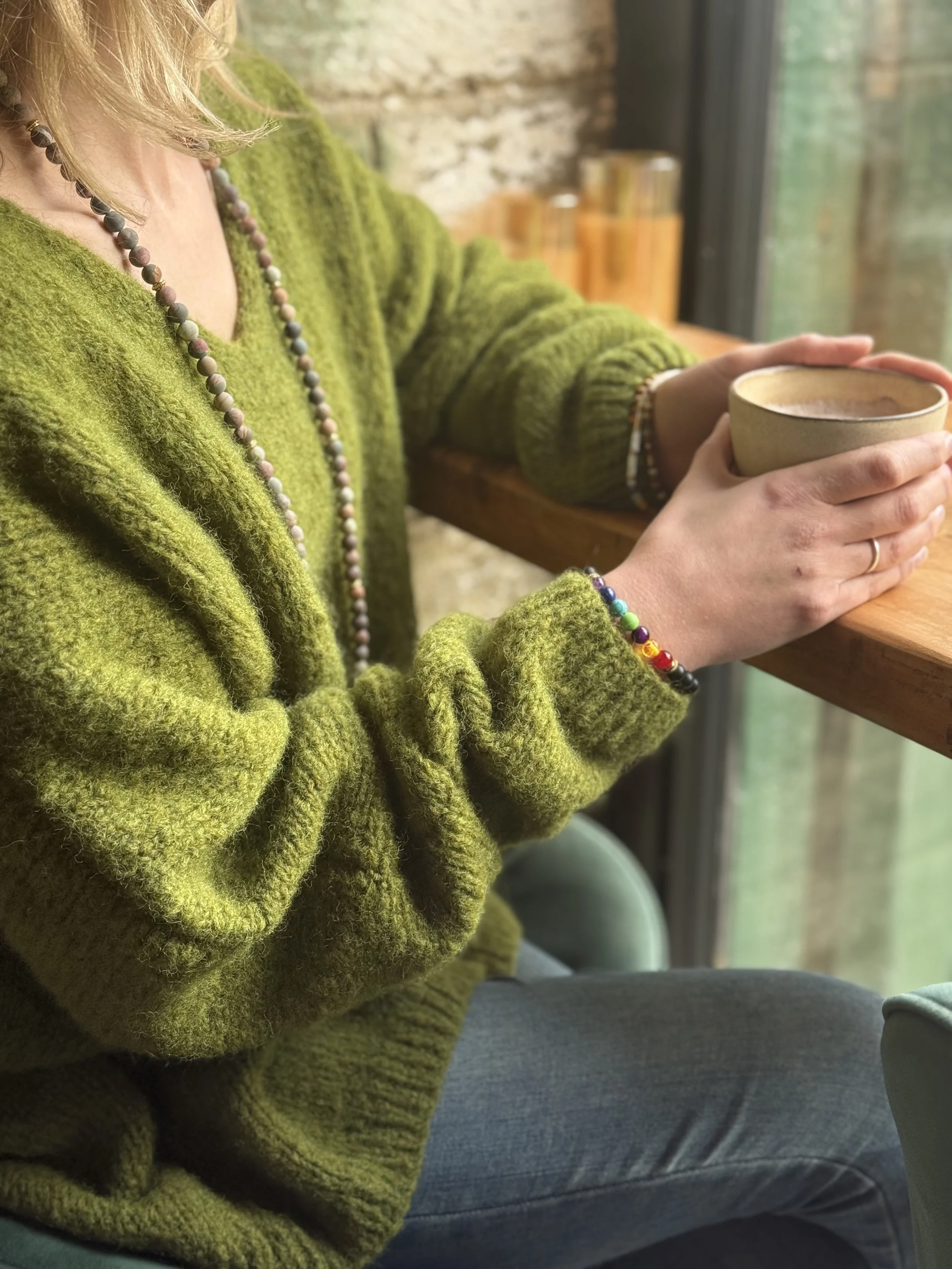 A person wearing a green fuzzy sweater and beaded jewelry sitting at a wooden counter, holding a ceramic mug near a large window with a ledge and a view of green trees outside.