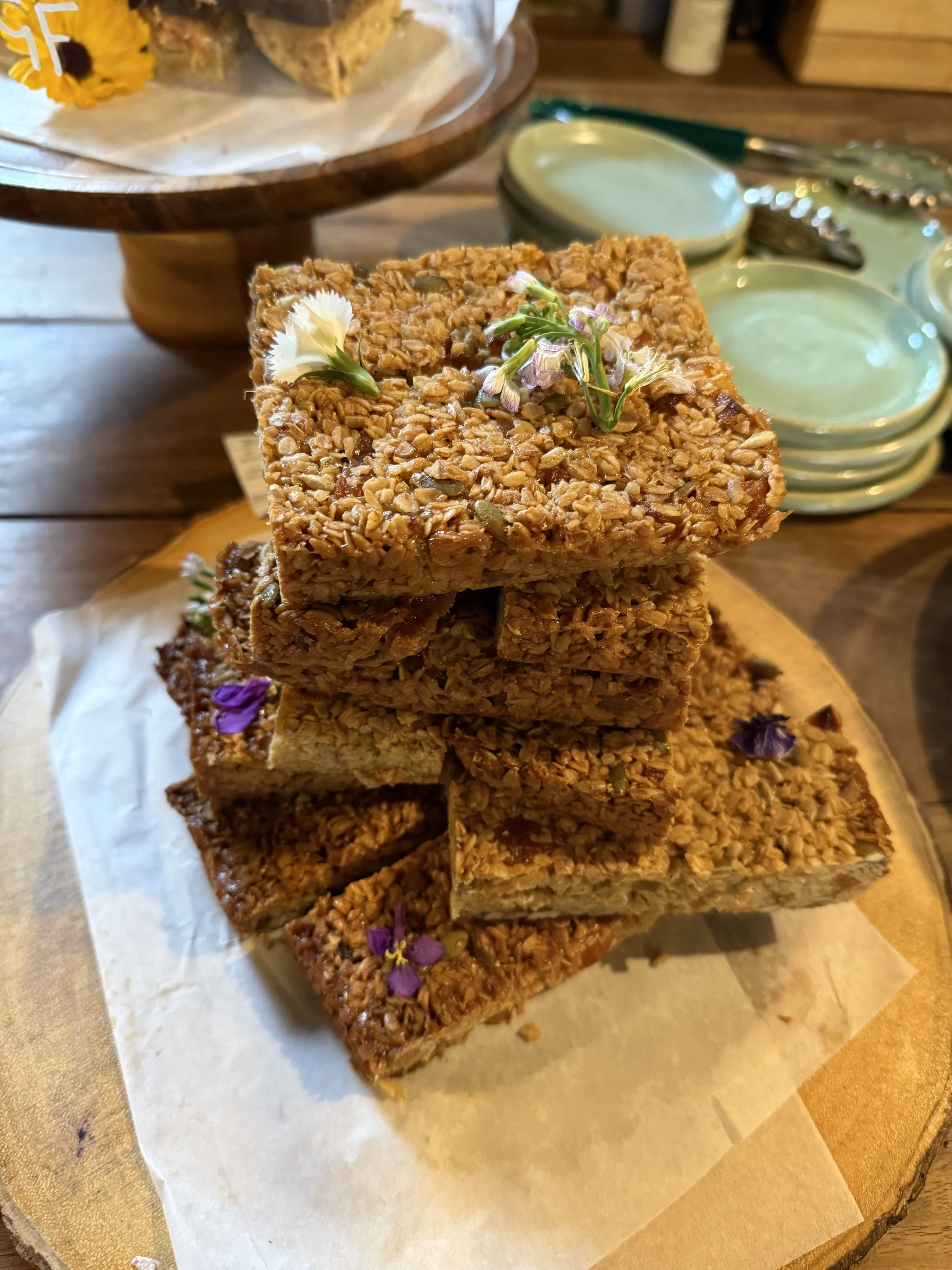 Stacked granola bars on a wooden board, decorated with purple and white edible flowers. In the background, there are small bowls and a spoon on a table, with a cake on a stand visible in the top left corner.