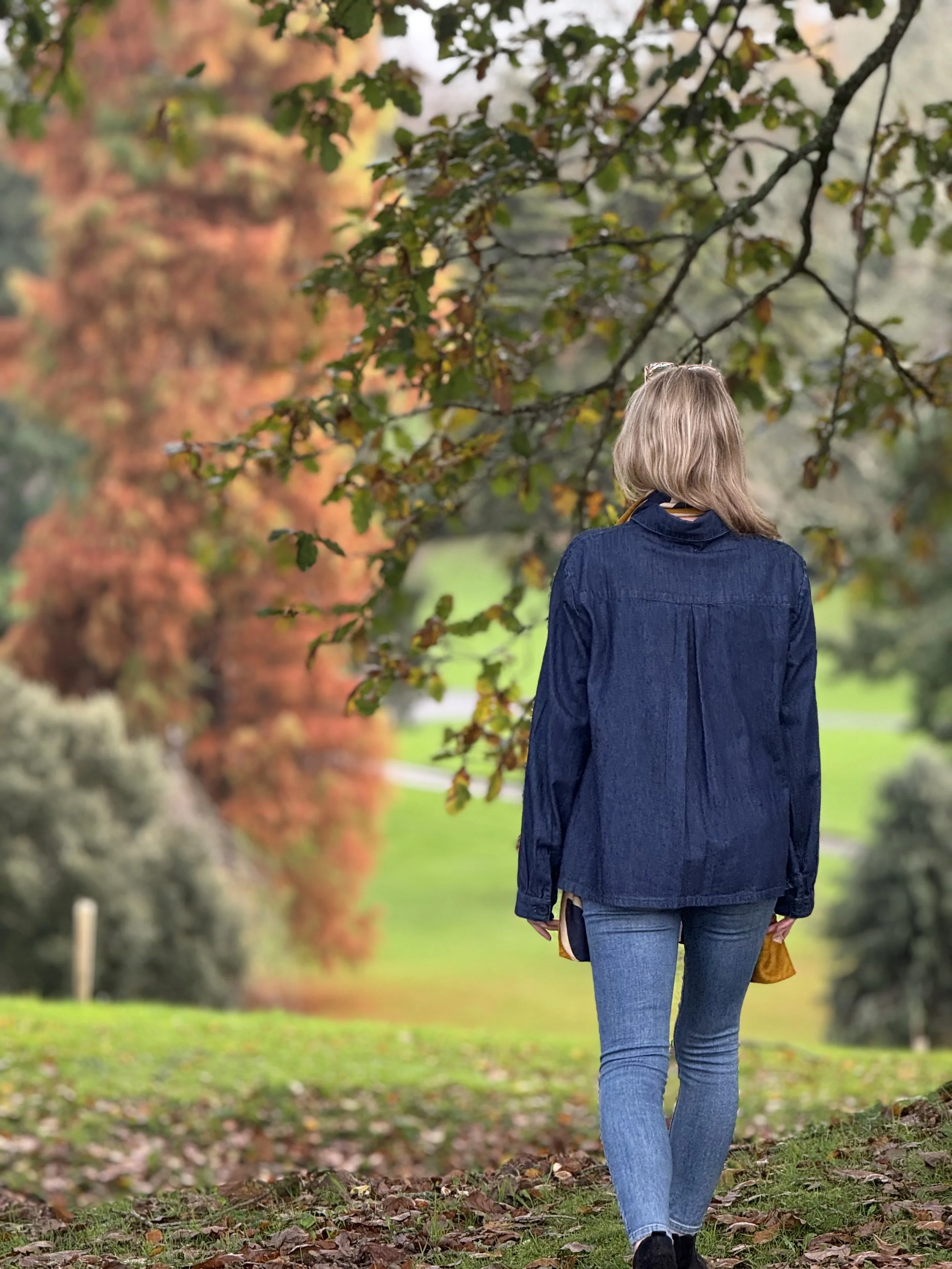 A woman walking outdoors on a path covered with fallen leaves in autumn. She is wearing a denim jacket and jeans, holding a yellow bag, with her back to the camera, surrounded by colorful autumn trees.