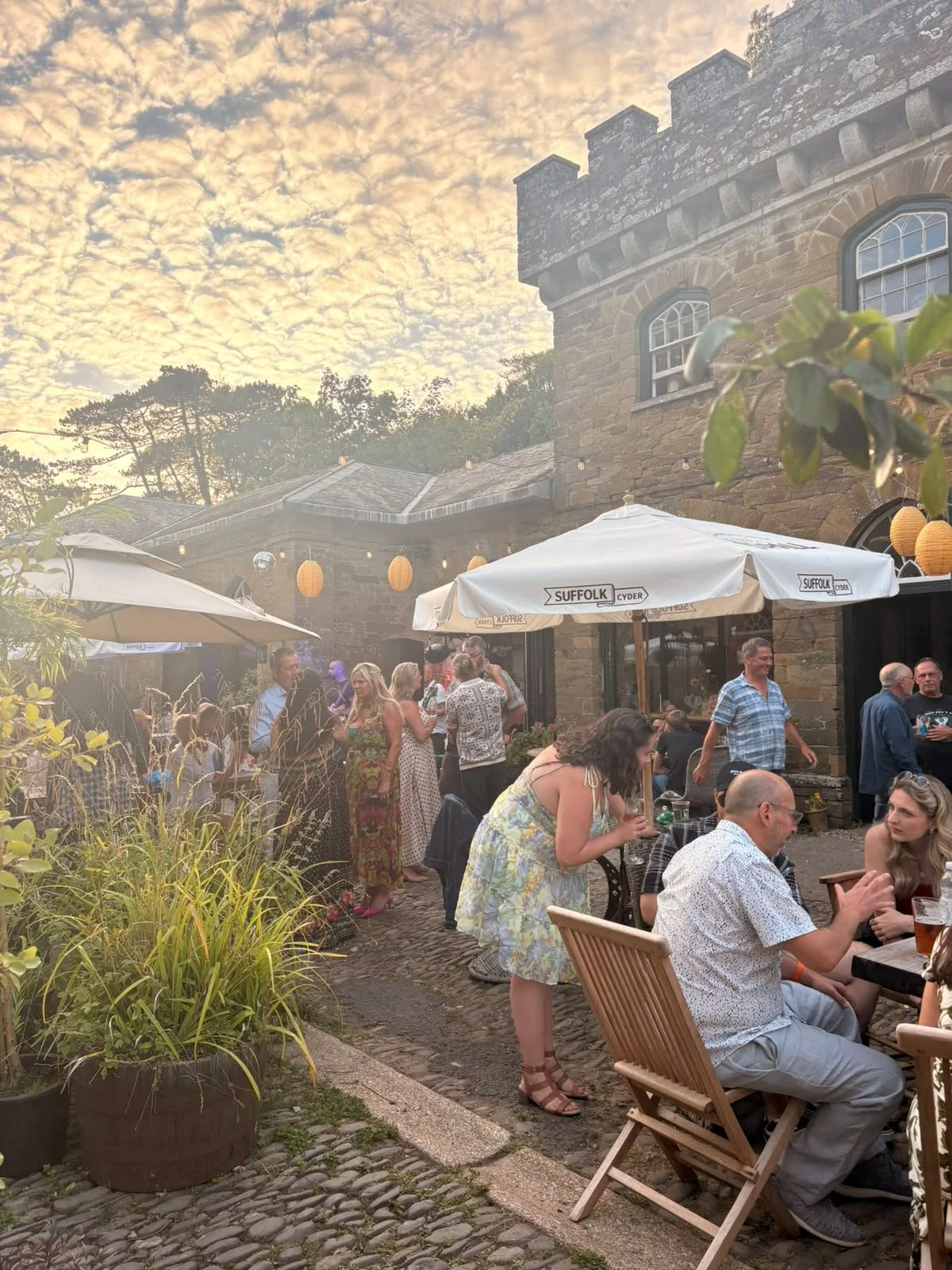 People gathered outside in a courtyard area during sunset, with some sitting at tables and others socializing, under umbrellas with 'Suffolk' branding, near a stone building with a castle-like roof, decorated with yellow lanterns and surrounded by gr