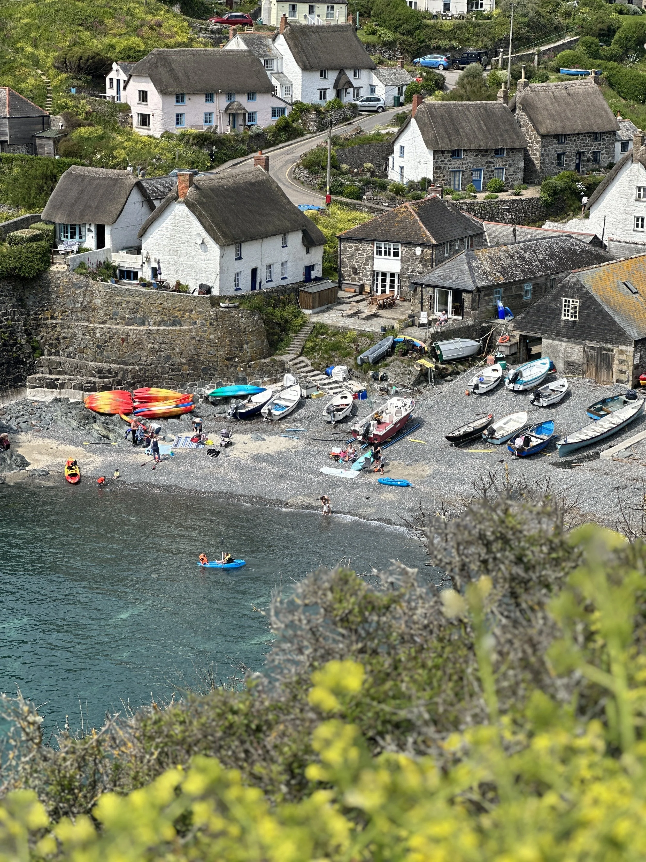 A small harbor with several boats and kayaks on a rocky beach. In the background, there are houses with thatched and shingled roofs on a hillside, surrounded by greenery.