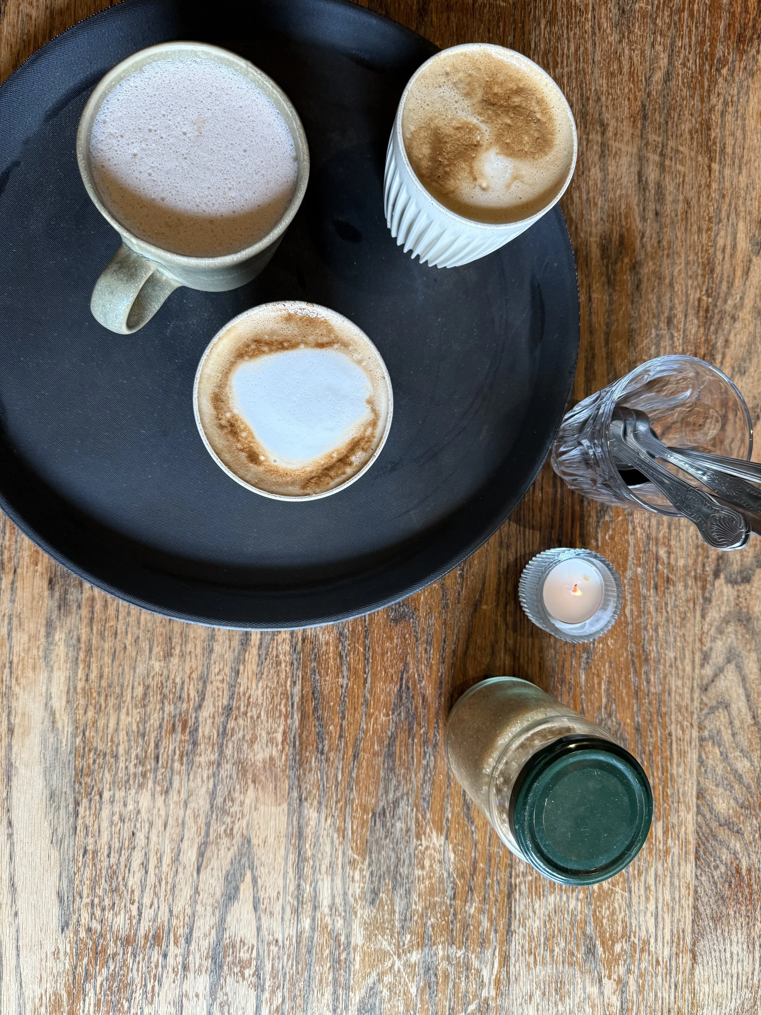 Three cups of coffee on a black tray, a glass of water with spoons, a small candle, and a jar on a wooden table.