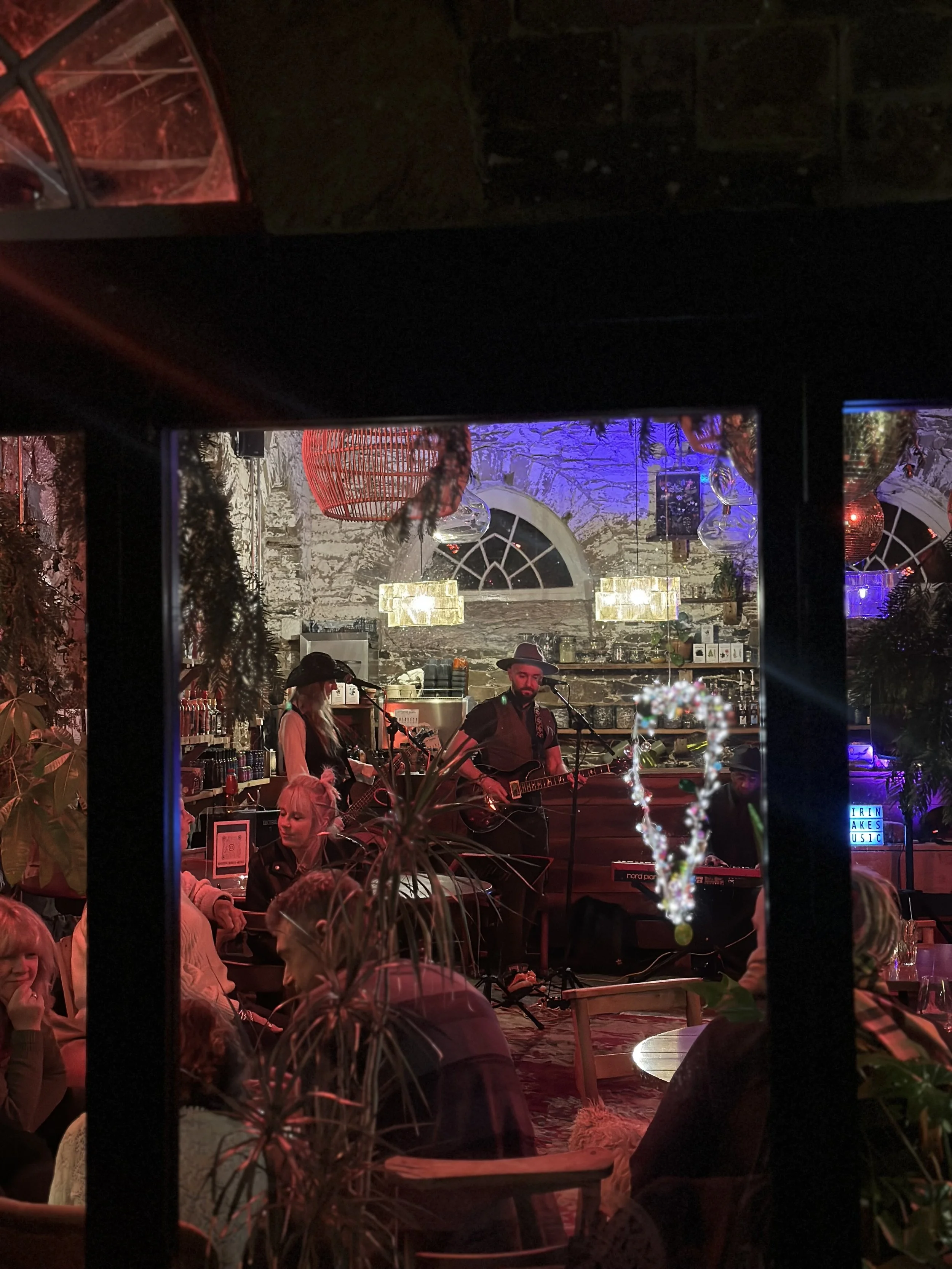 A band performing live music in a cozy, dimly lit restaurant or bar with exposed brick walls, colorful lights, and an audience enjoying the show.