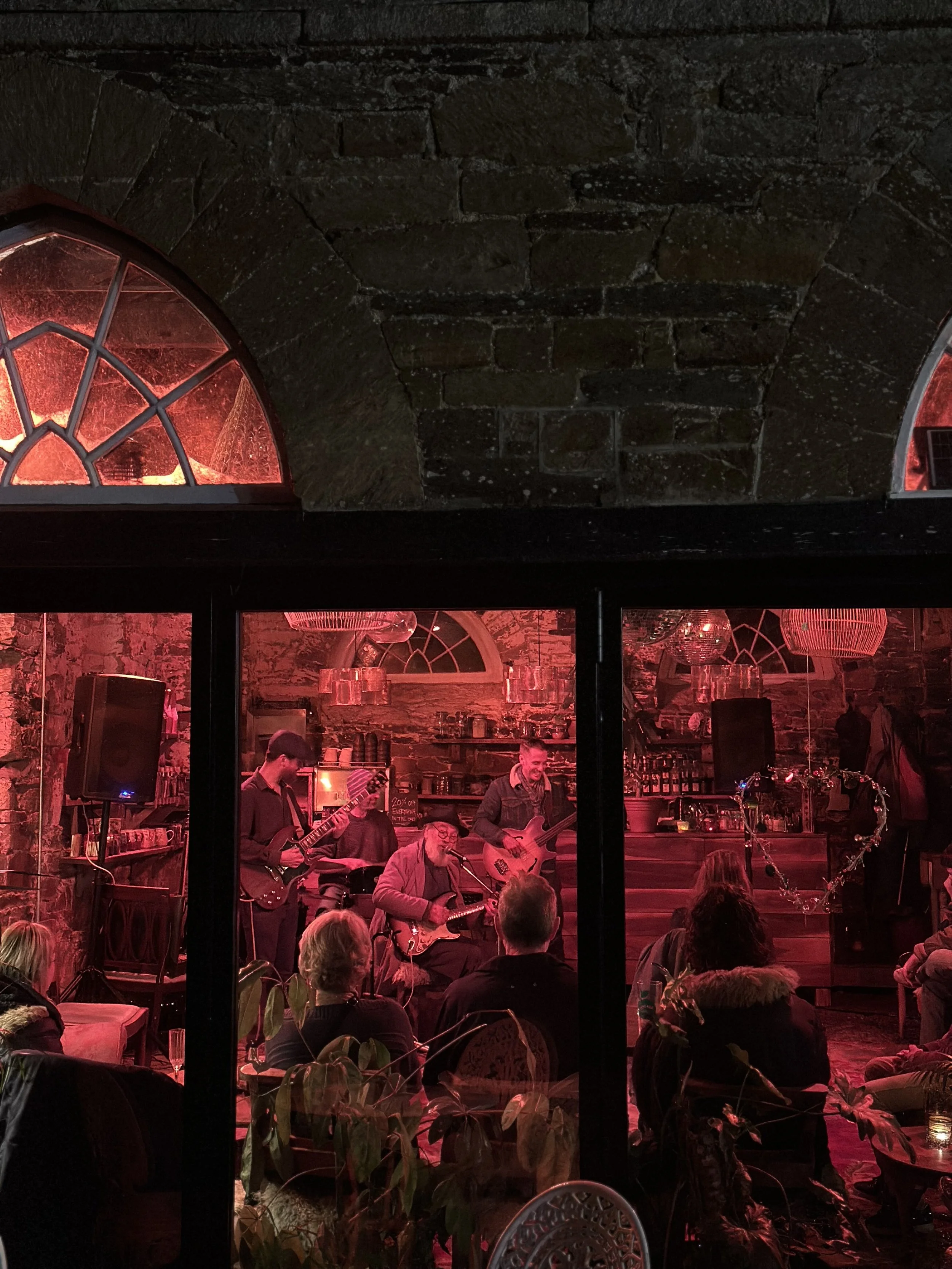 A live music performance with a band playing on stage, visible through a window, in a cozy, brick-walled venue with red lighting and an audience watching.