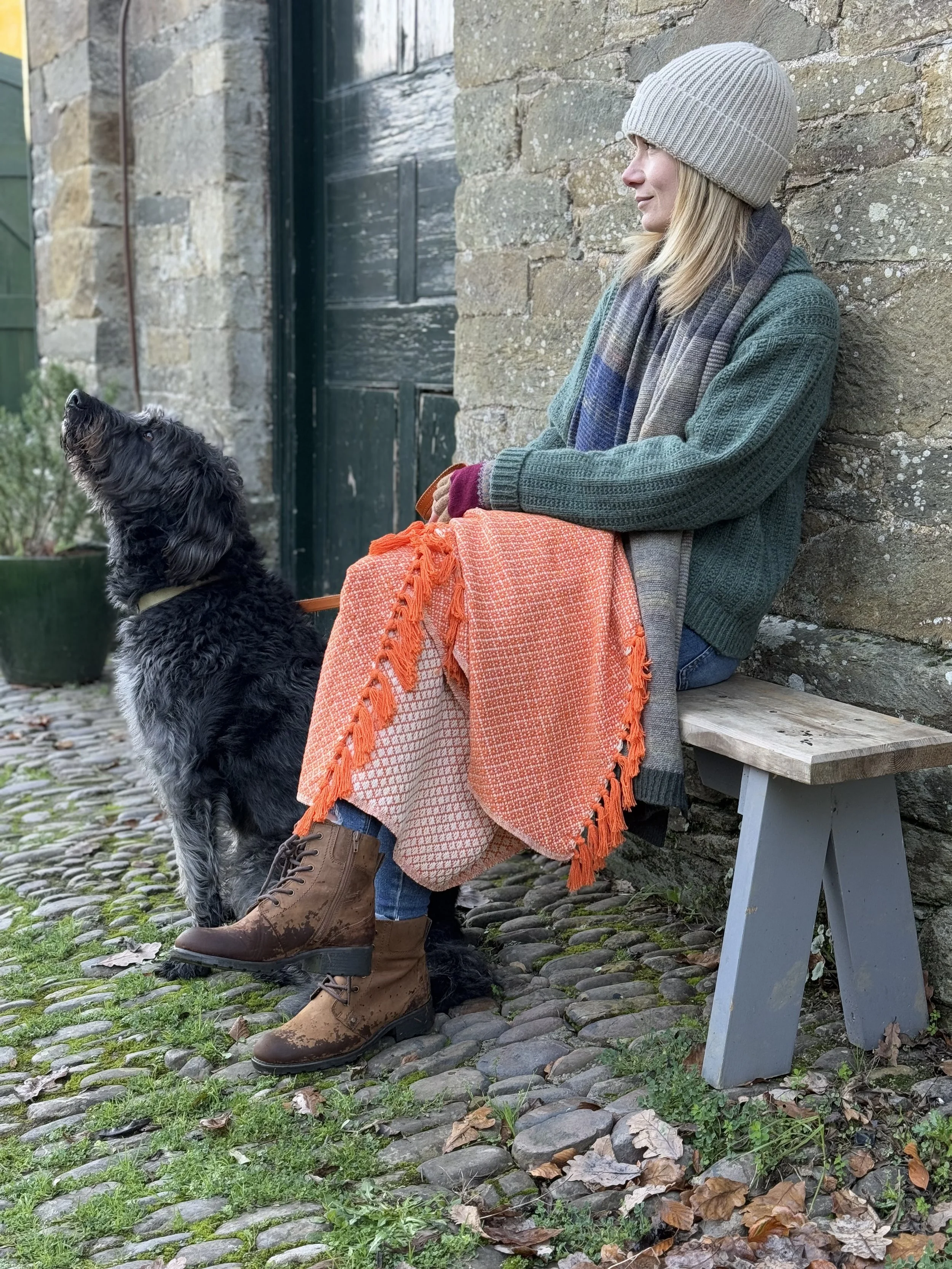 A woman with blonde hair, wearing a beige beanie, a green sweater, a multicolored scarf, brown hiking boots, and jeans, is sitting on a wooden bench against a stone wall. Next to her is a black and gray curly-haired dog sitting on cobblestone ground 
