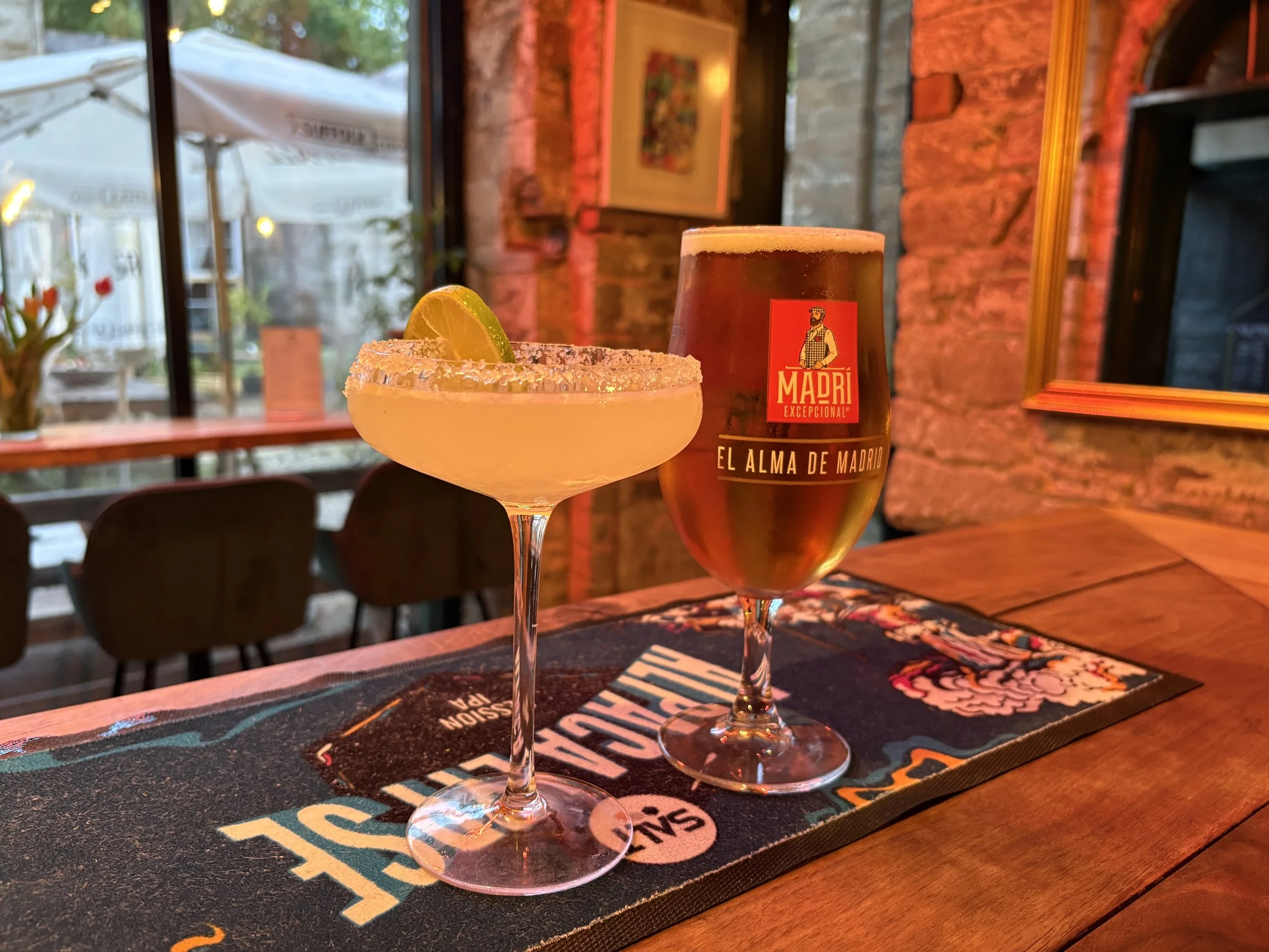 A cocktail glass with a salted rim, garnished with a lemon wedge, and a pint glass of beer on a wooden table in a bar or restaurant setting.