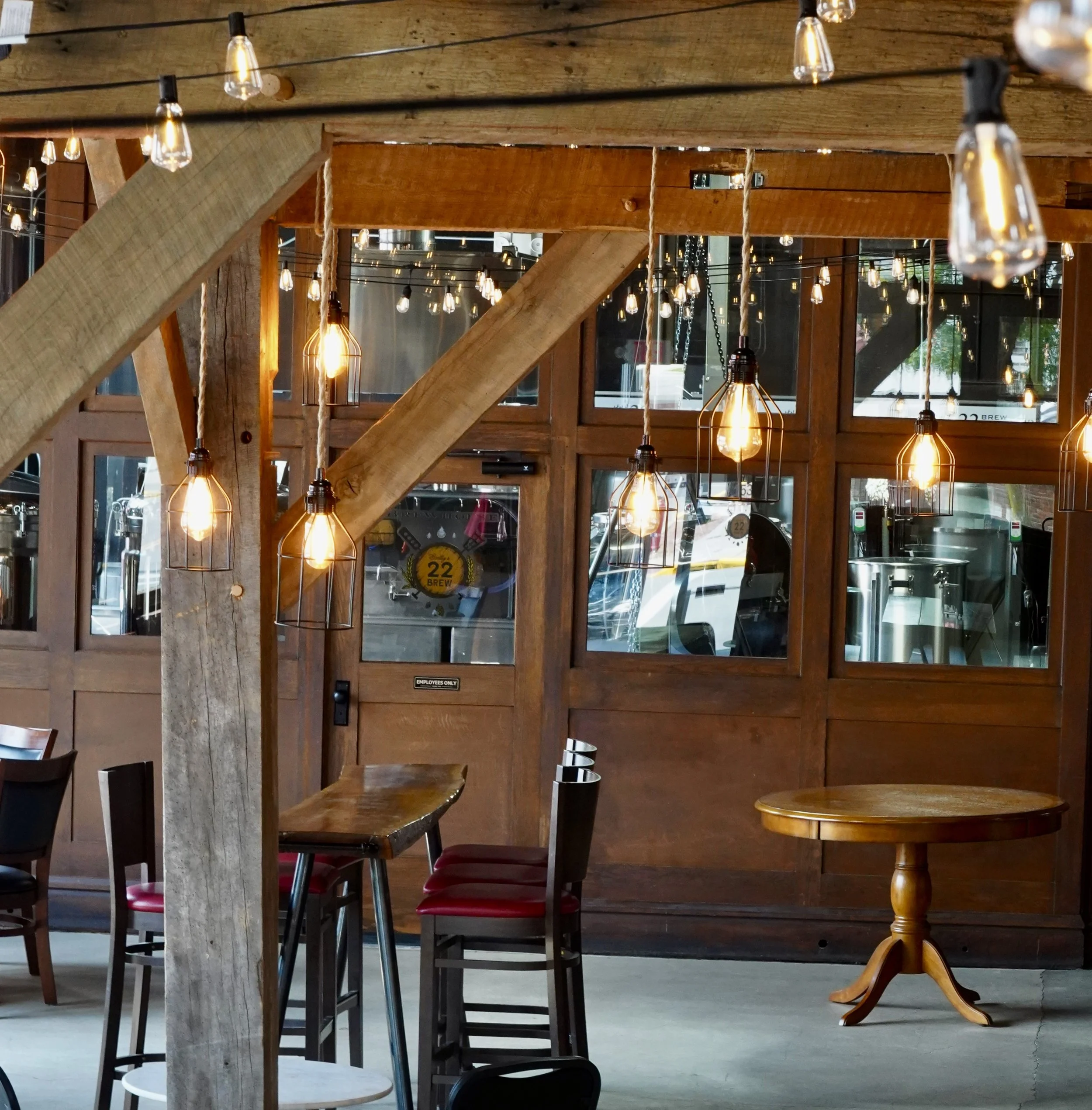 Interior of a cozy restaurant or cafe with wooden furniture, hanging Edison light bulbs, and a large wooden window showing brewing equipment outside.