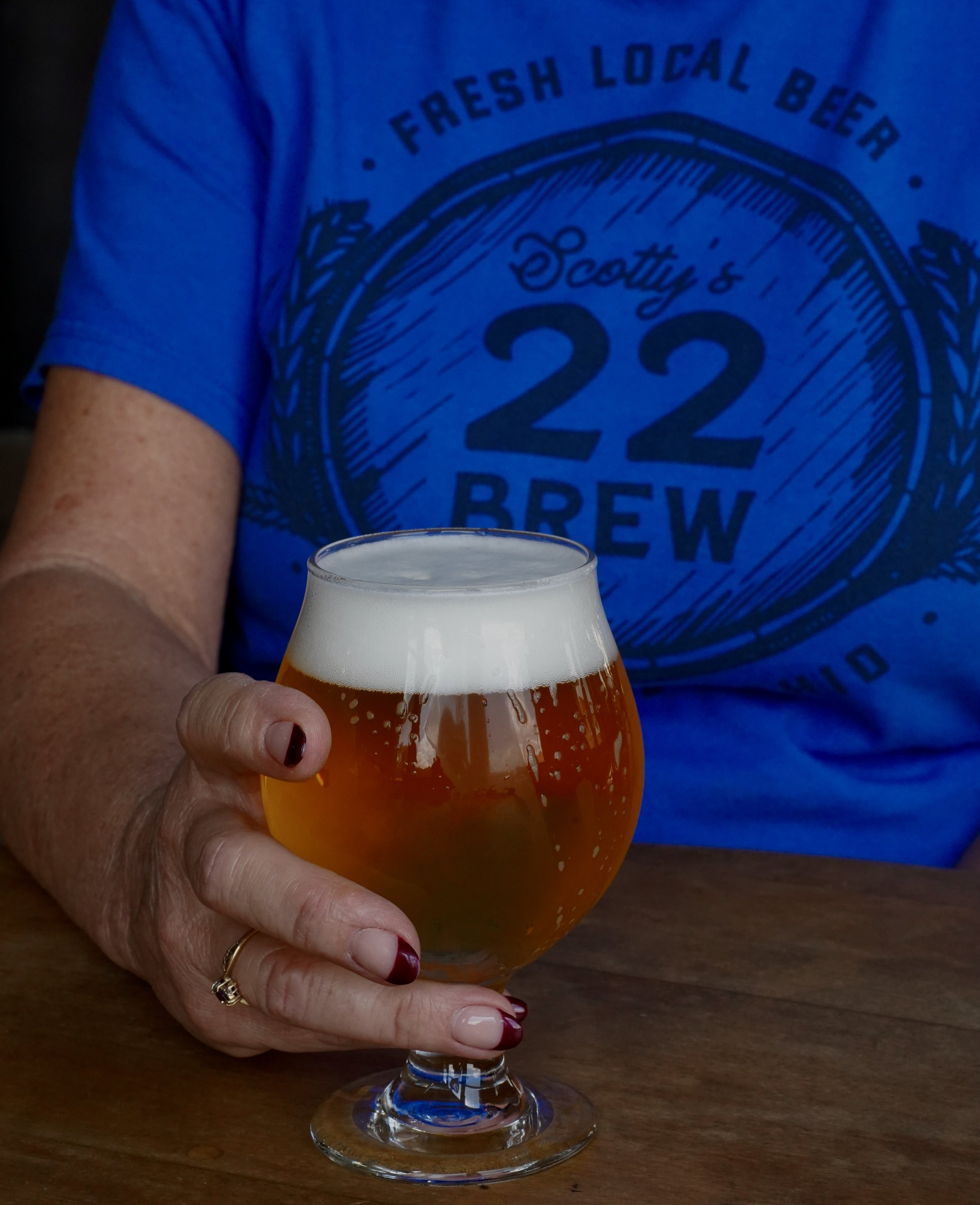 Person holding a glass of beer with a white foam head, wearing a blue T-shirt with a logo that says Scottie's 22 Brew.