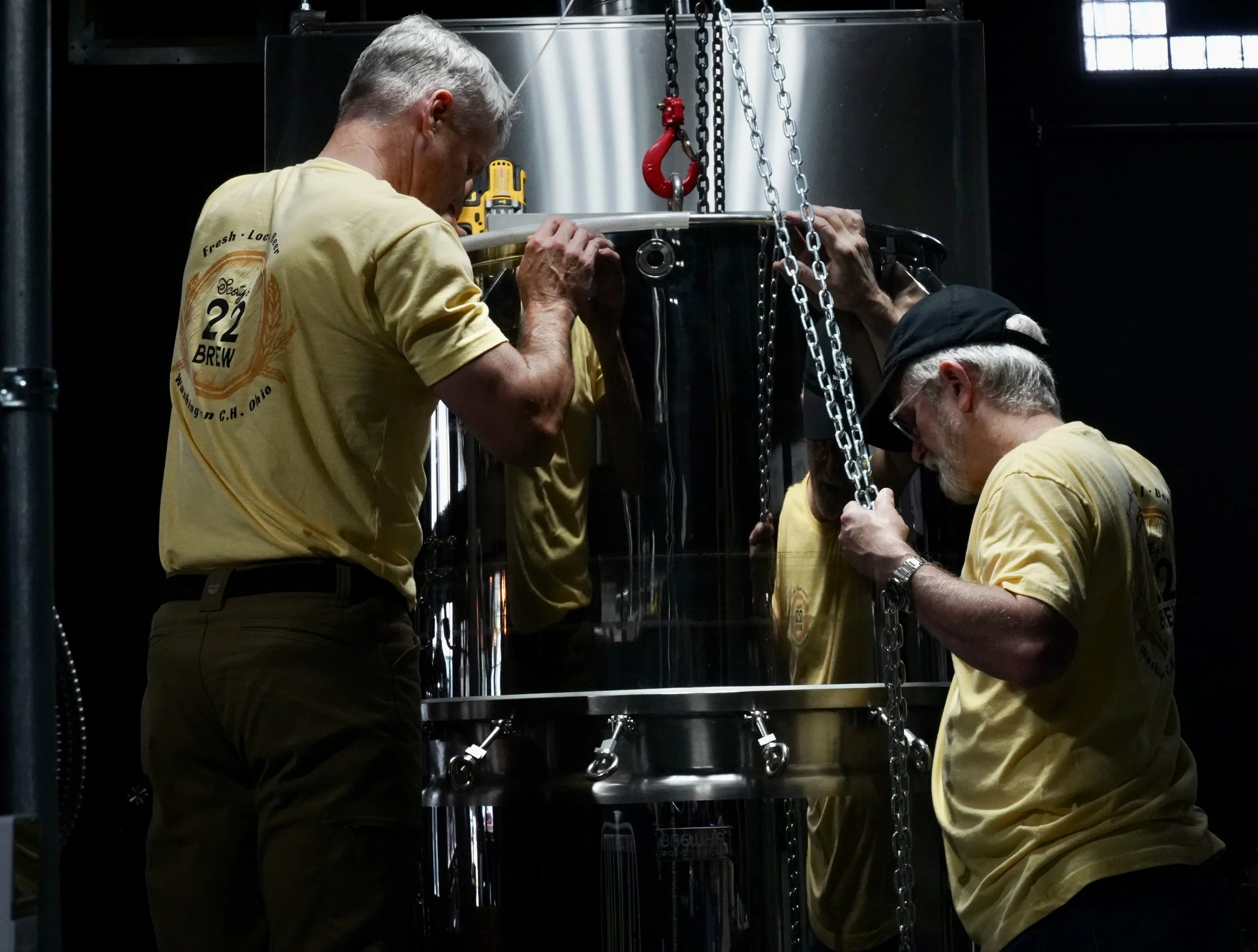 Two men wearing yellow shirts working together on a large metallic brewing vessel, inspecting or repairing it, in a dimly lit industrial setting.