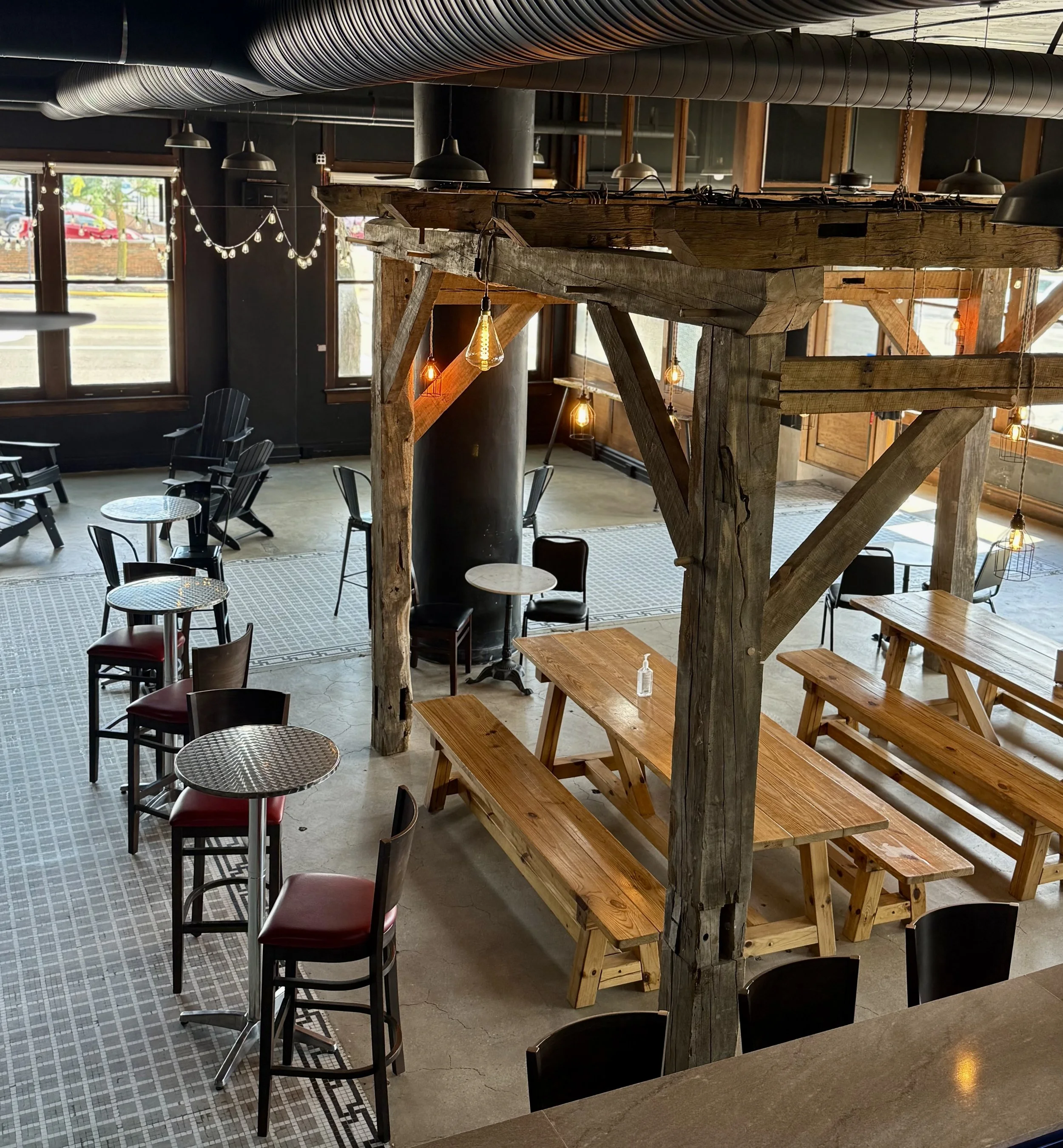 Interior of a rustic-style cafe with wooden beams, string lights, high-top tables with red cushioned chairs, and outdoor seating visible through large windows.