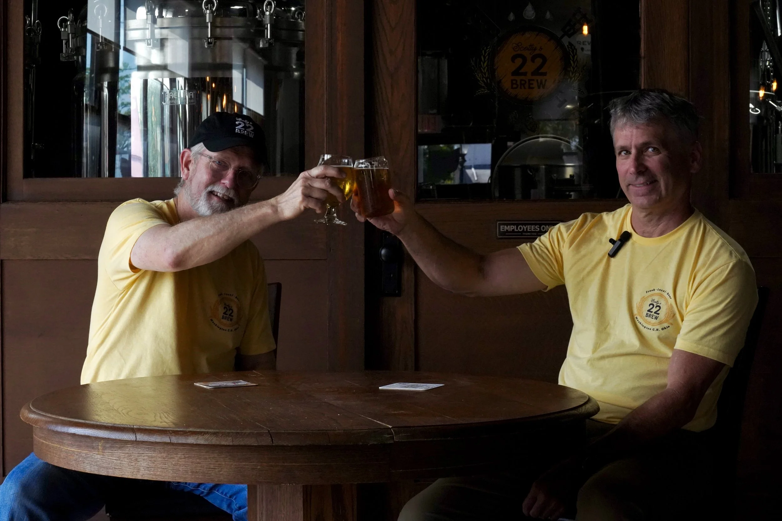 Two men sitting at a wooden table, wearing yellow t-shirts with a '22 Brew' logo, toasting with glasses of beer inside a brewery, with brewing equipment visible in the background.