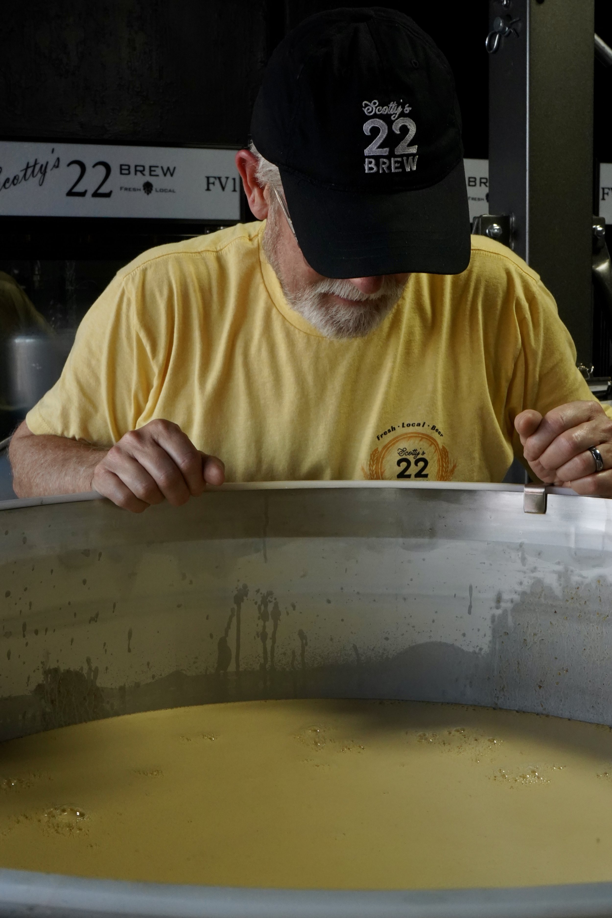 A man with a white beard and glasses, wearing a black cap with 'Scotty's 22 Brew' logo and a yellow shirt with the same logo, leaning over a large metal container filled with a yellow liquid.