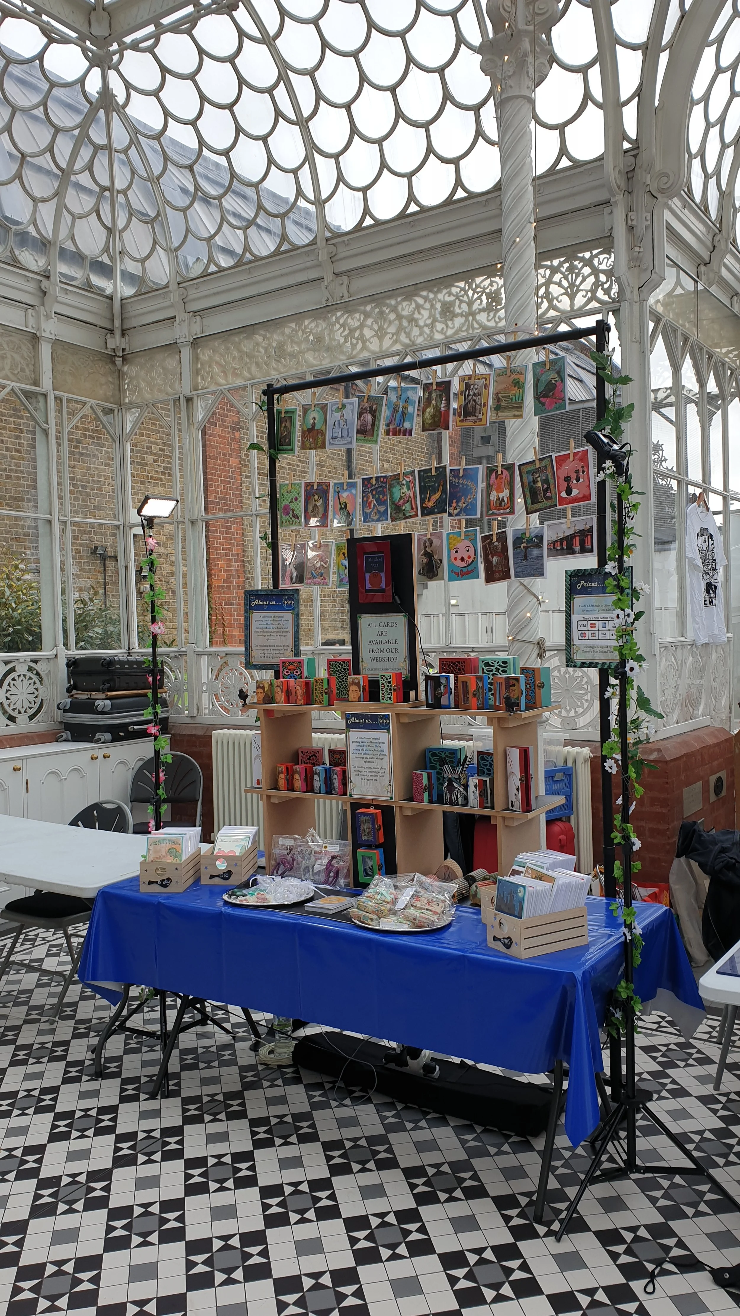 A display table and stand with various colorful arts, cards, and greeting cards inside a glass-domed pavilion with patterned floor
