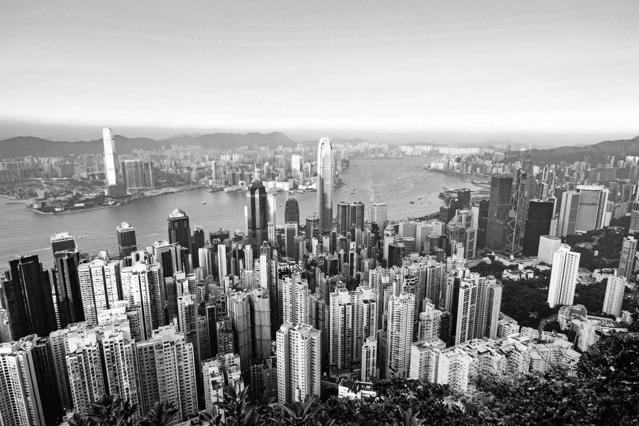 Black and white aerial view of a dense city skyline with numerous high-rise buildings, a river, and mountains in the background.